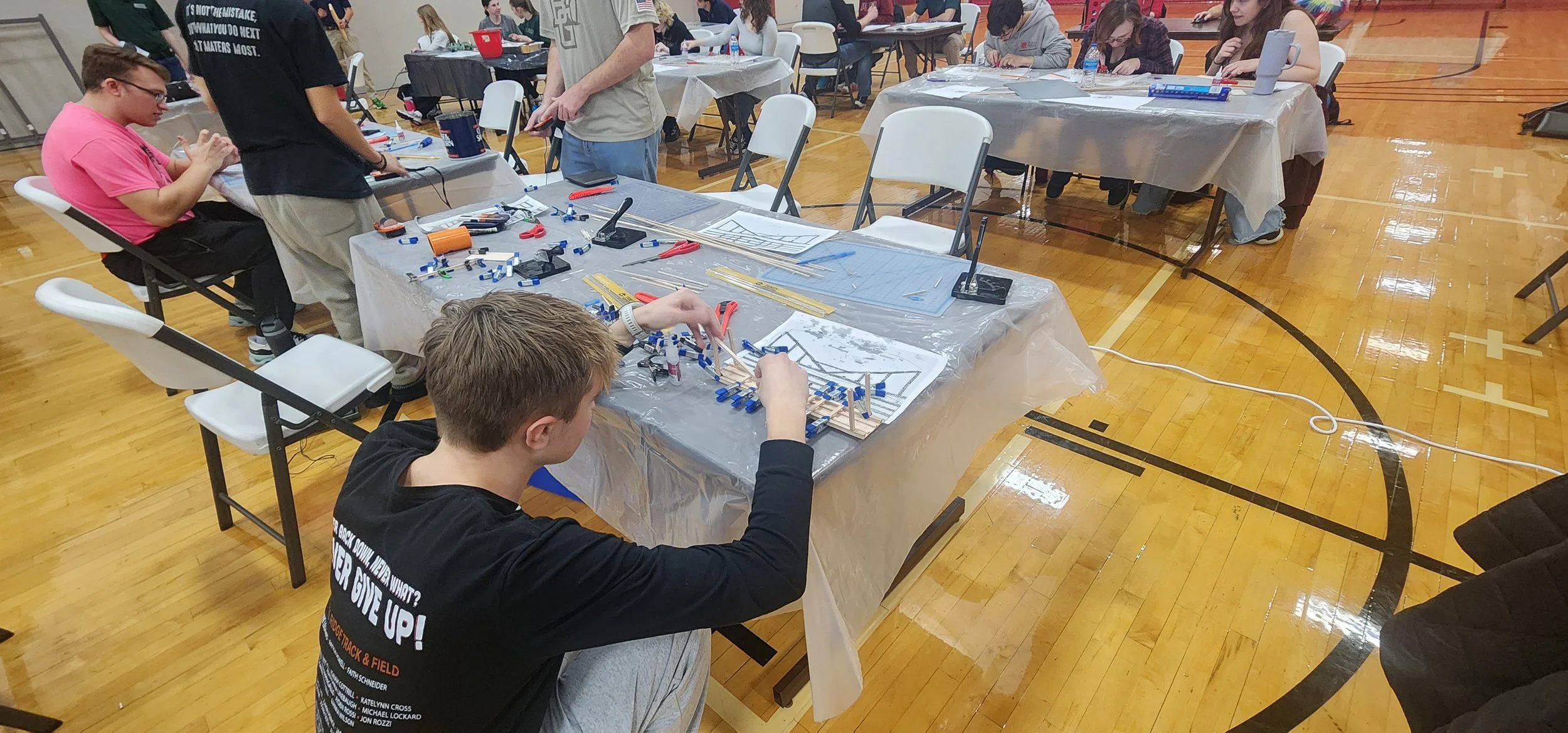 A group of people working on a robotics project at tables covered with plastic tablecloths in a gymnasium. They are building structures with wooden sticks and blue plastic pieces, with blueprints and tools scattered around.