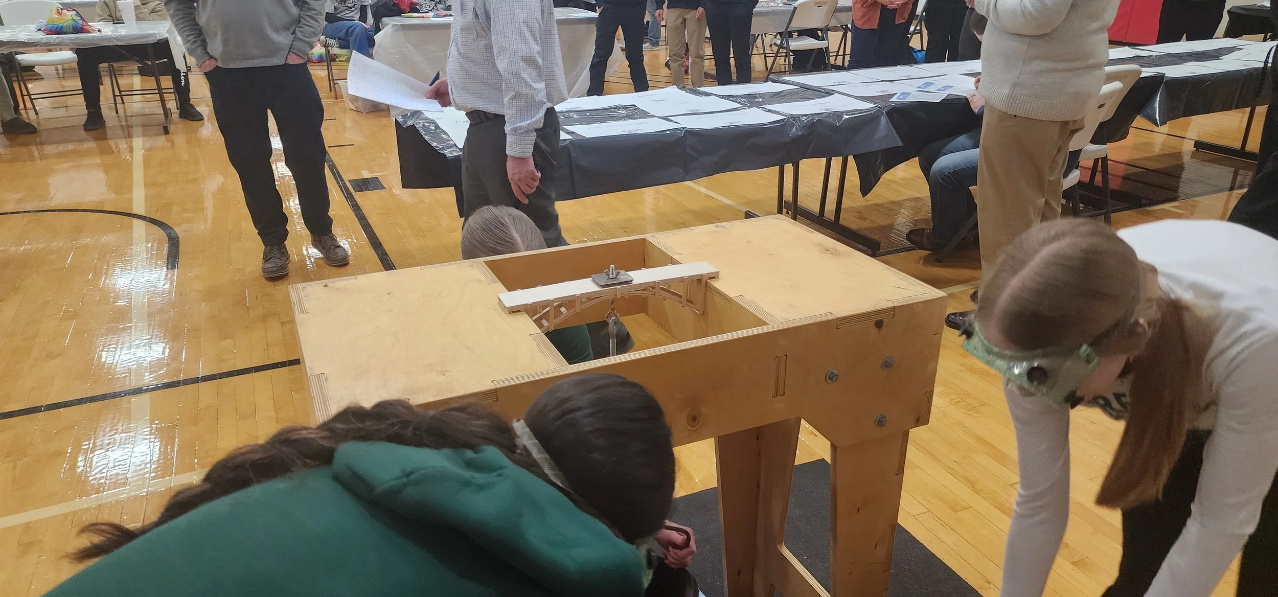 A girl is crouching down to look through a hole in a wooden structure during an event at a gymnasium. Several people are standing around, some are watching or taking notes, and tables are lined up with papers and displays. The floor is wooden, and th
