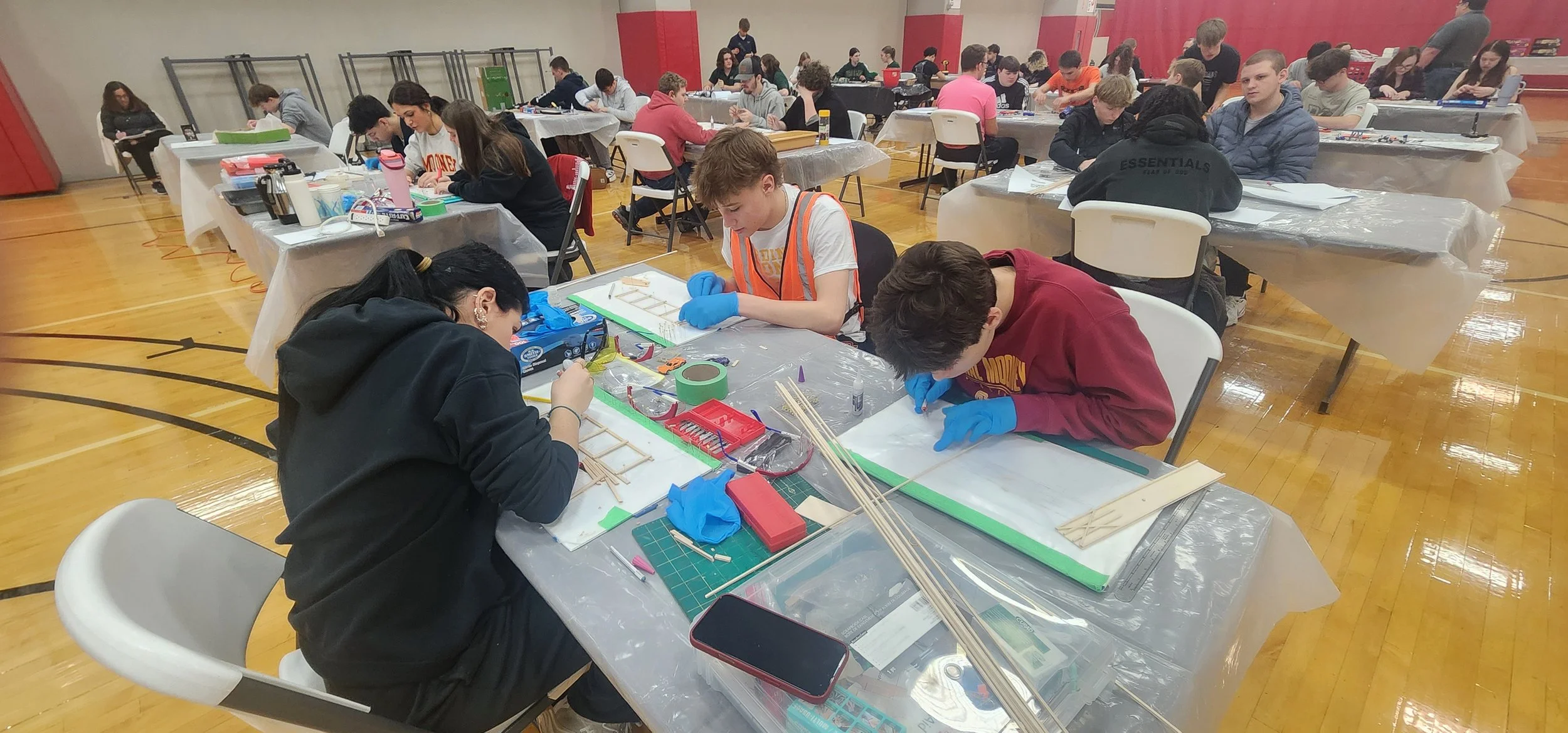 Students working on model-building projects at tables covered with tools and materials in a gymnasium.