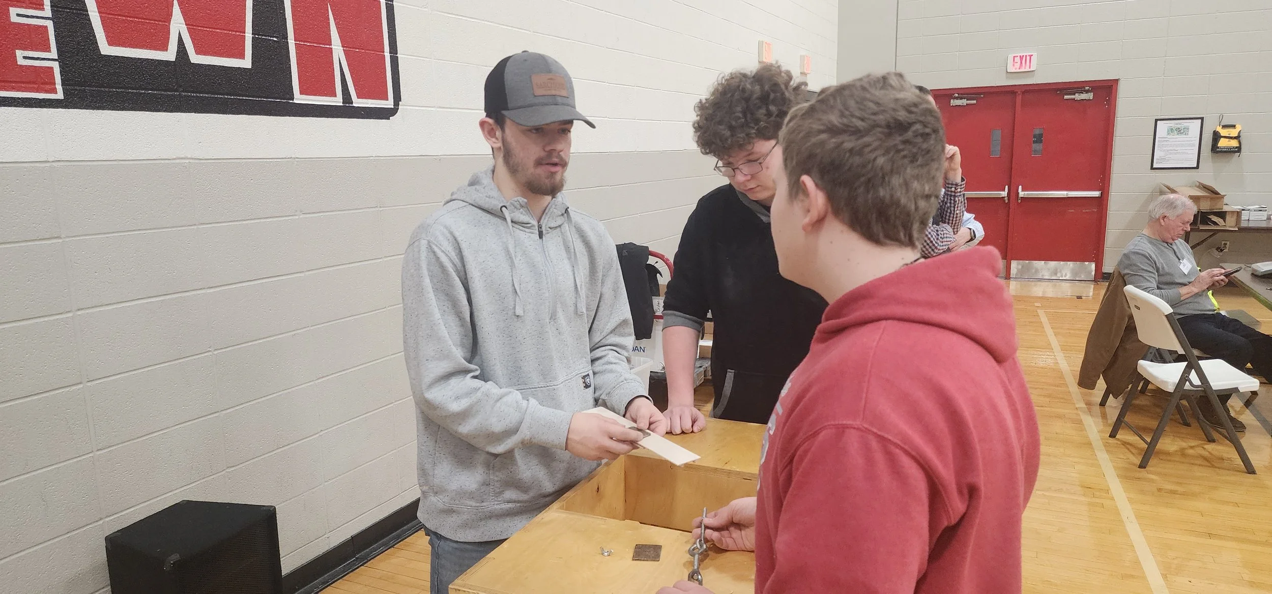 Three young men are at a table, one holding a paper and the others looking at it, with an older man sitting in the background using his phone in a gymnasium.