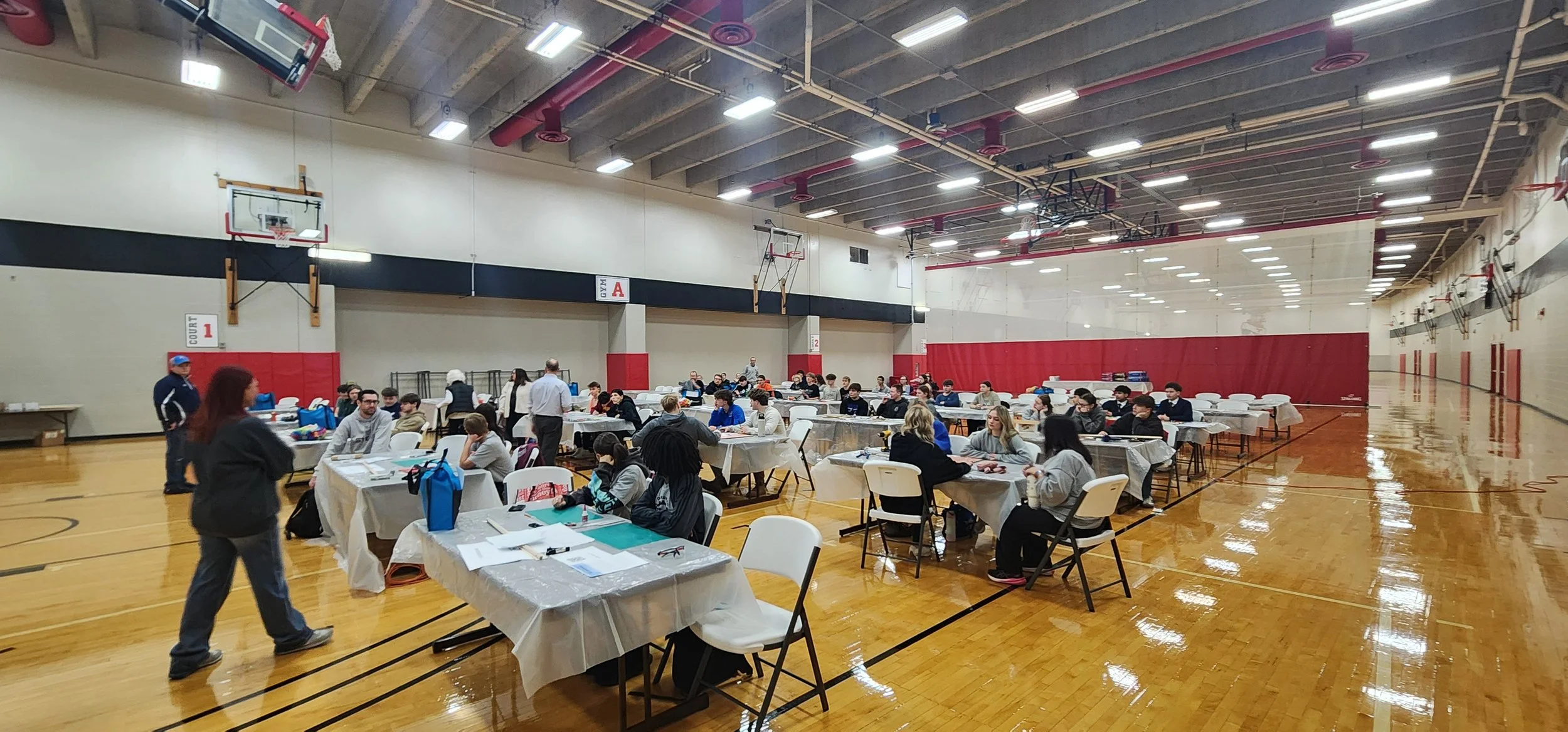 People sitting at tables in a gymnasium, participating in an event or competition, with some standing and walking around. The gym has basketball hoops and red padding on the walls.