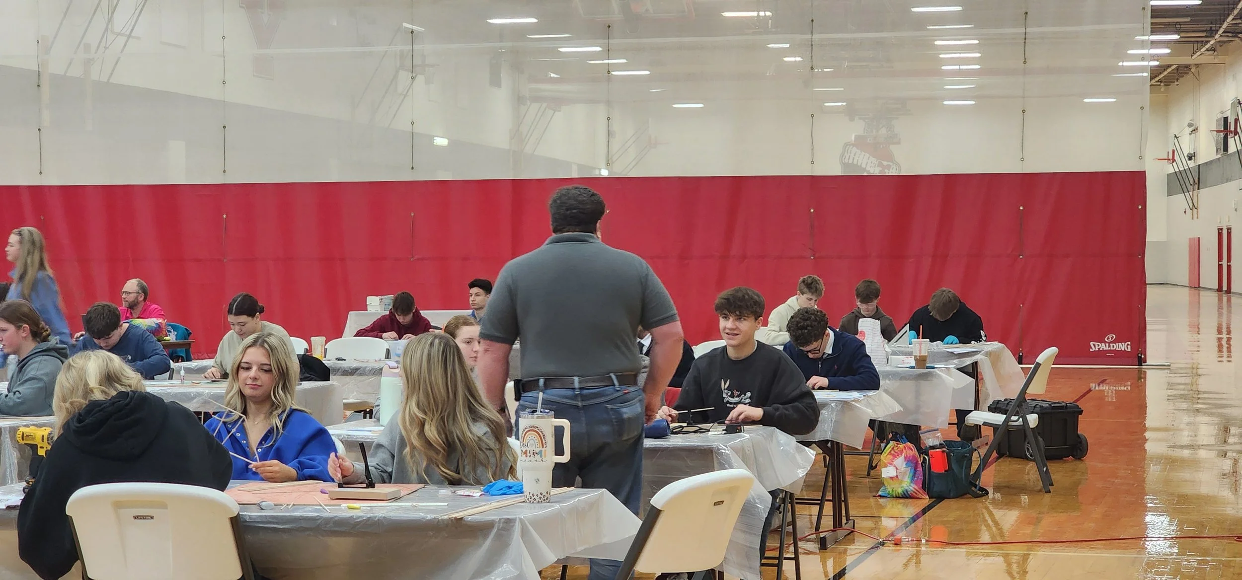 A group of teenagers and young adults seated at tables in a gymnasium, with some working on electronic devices and others talking, under the supervision of an adult.