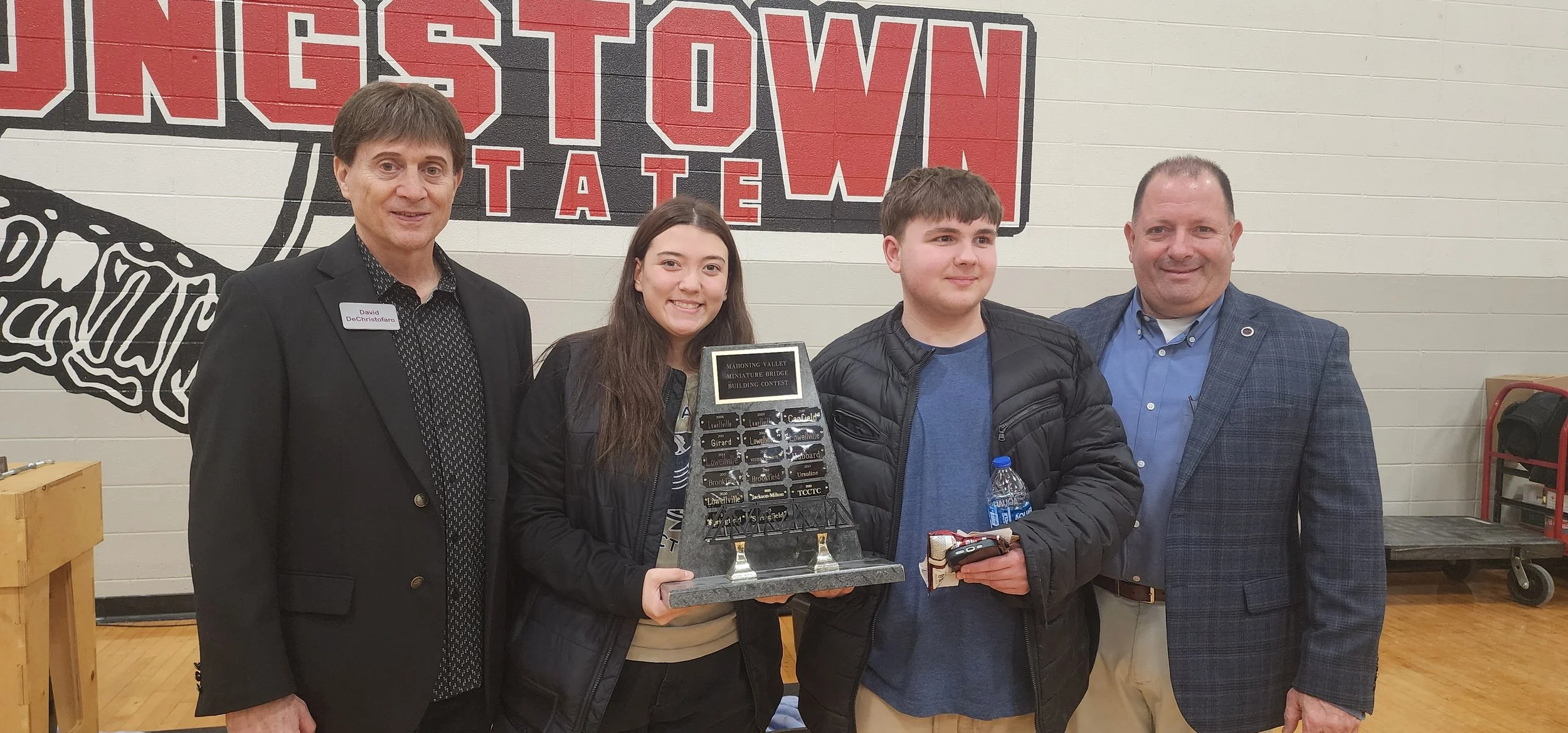 Four people standing in front of a wall with a large sign reading "TOWN" and "TATE". The person second from the left holds a display plaque, and the person second from the right holds a water bottle and car keys. The group appears to be at a school o