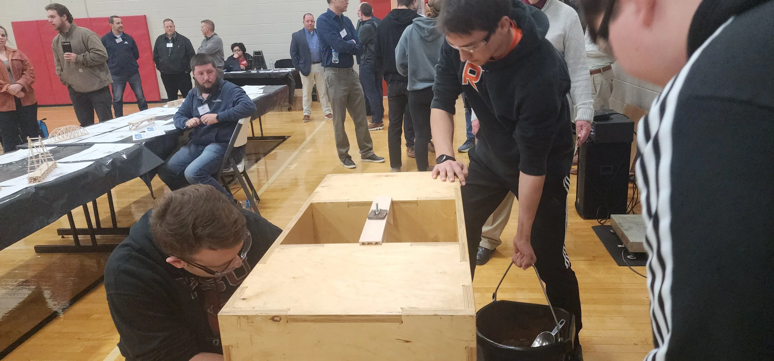 People participating in a science or engineering activity at an indoor event, with some working on a wooden structure and others observing, in a gymnasium.