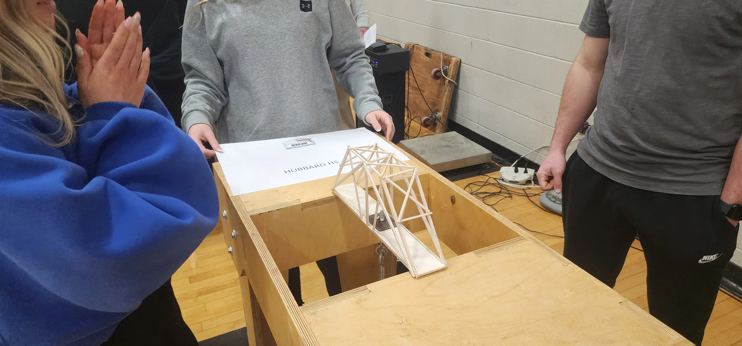 Students observe a wooden bridge model and a blueprint labeled 'Hubbard HS,' on a table at a classroom or workshop.
