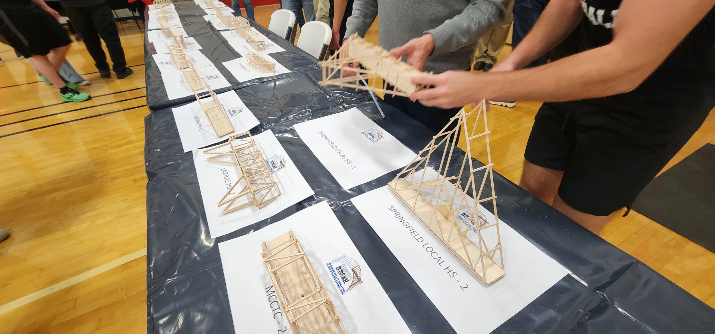 People at a table with miniature wooden bridge models displayed on paper signs.