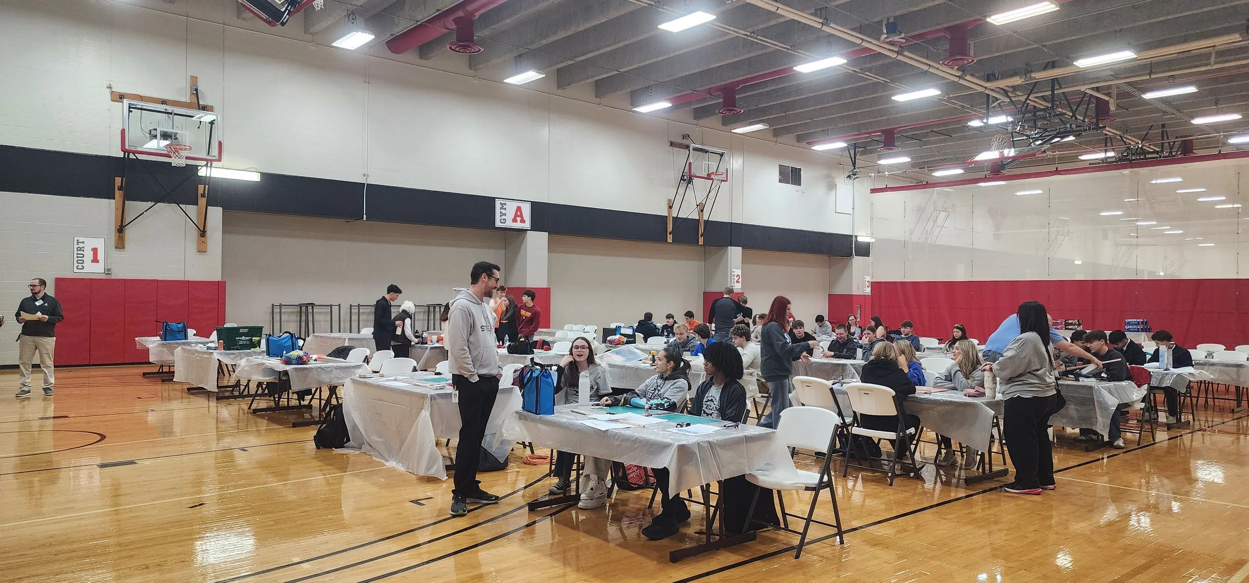A gymnasium turned into a voting or registration site with tables covered in plastic, people sitting and standing, and some officials managing the event.