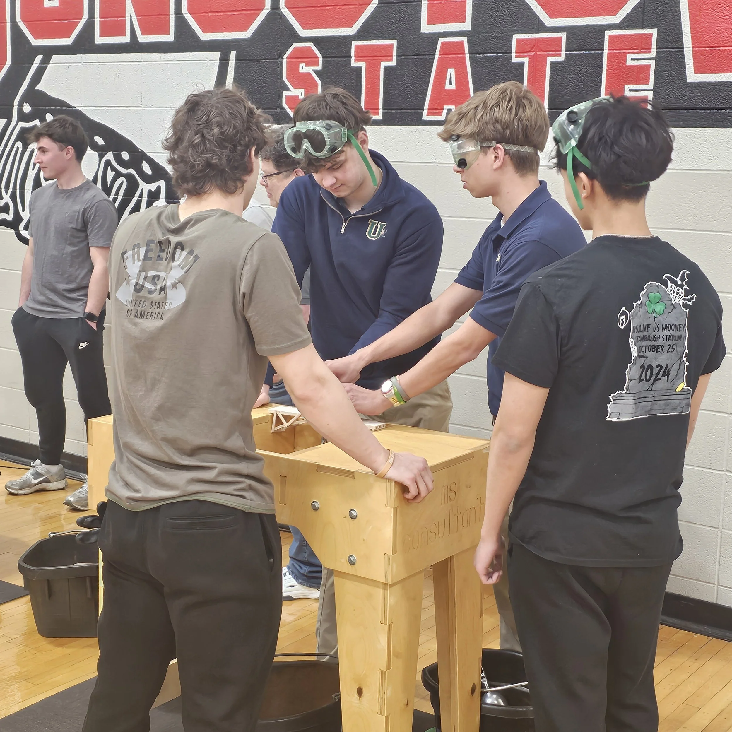 Group of young men and women gathered around a wooden table at an indoor event, some wearing protective goggles, with a large red and black sign reading 'STATE' in the background.
