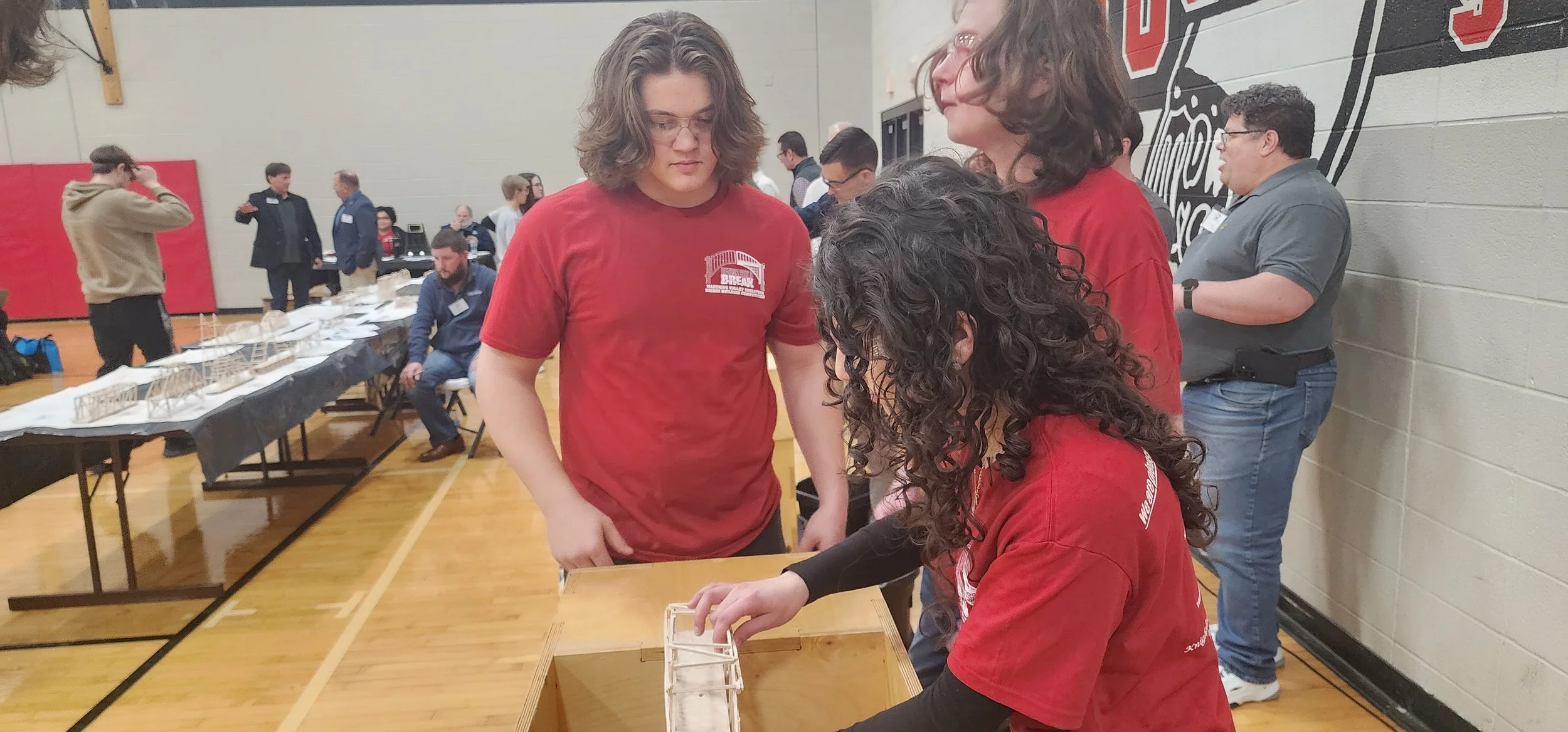 People participating in a bridge-building competition in a gymnasium. A young woman is attaching or adjusting a small bridge model made of wood and white supports on a table, while others watch and work nearby.