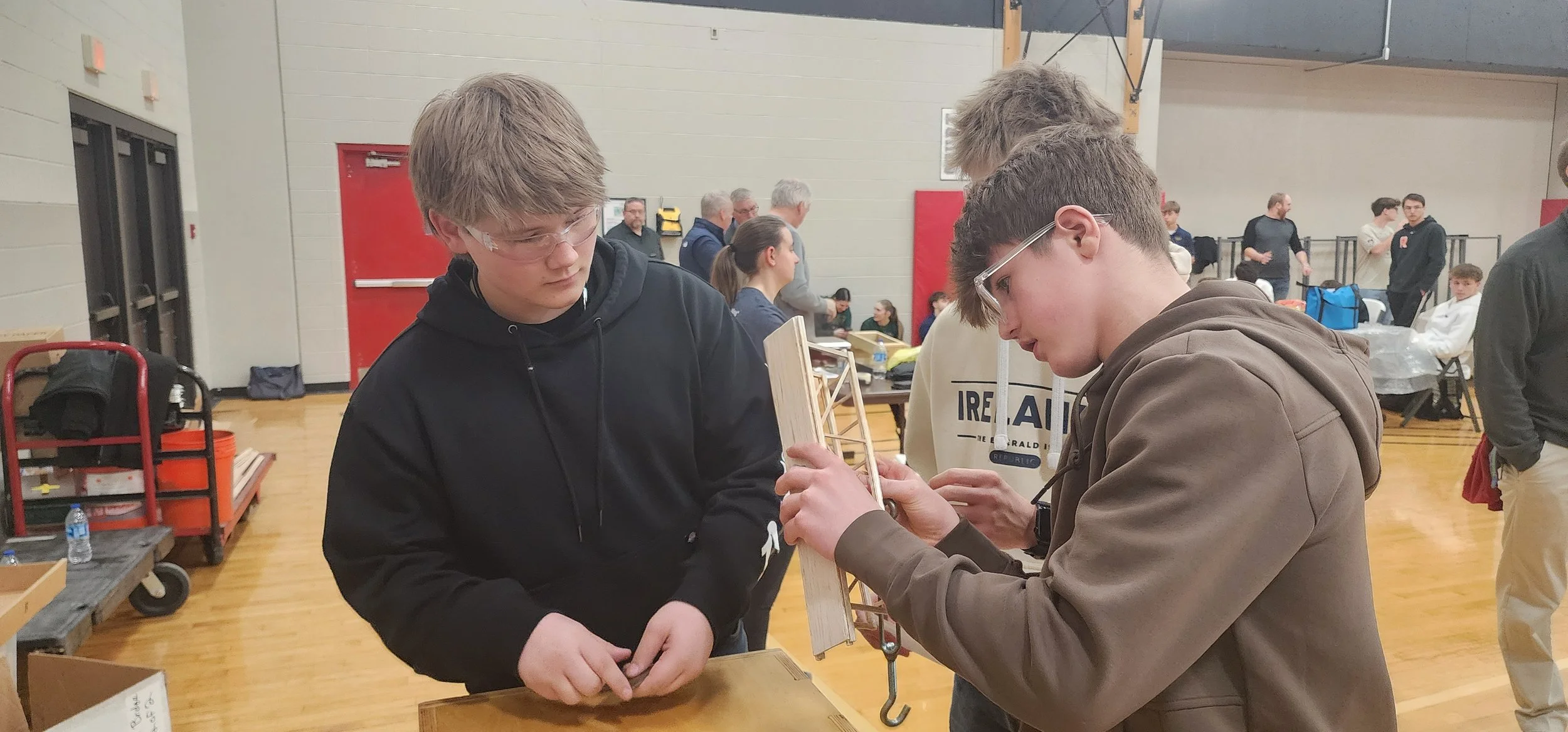 Two young men wearing glasses and hoodies are examining a wooden item at an indoor event in a gymnasium. Background shows other people, tables, and equipment.