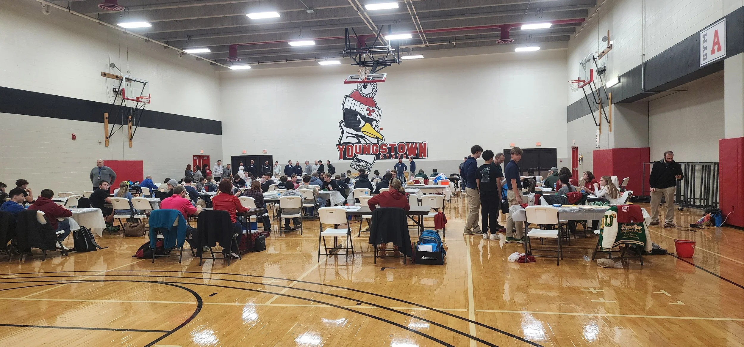 A gymnasium filled with students sitting at tables and talking, with some standing and walking around. The walls have a large logo with a duck wearing sunglasses and the words "Youngstown State" on the back wall.