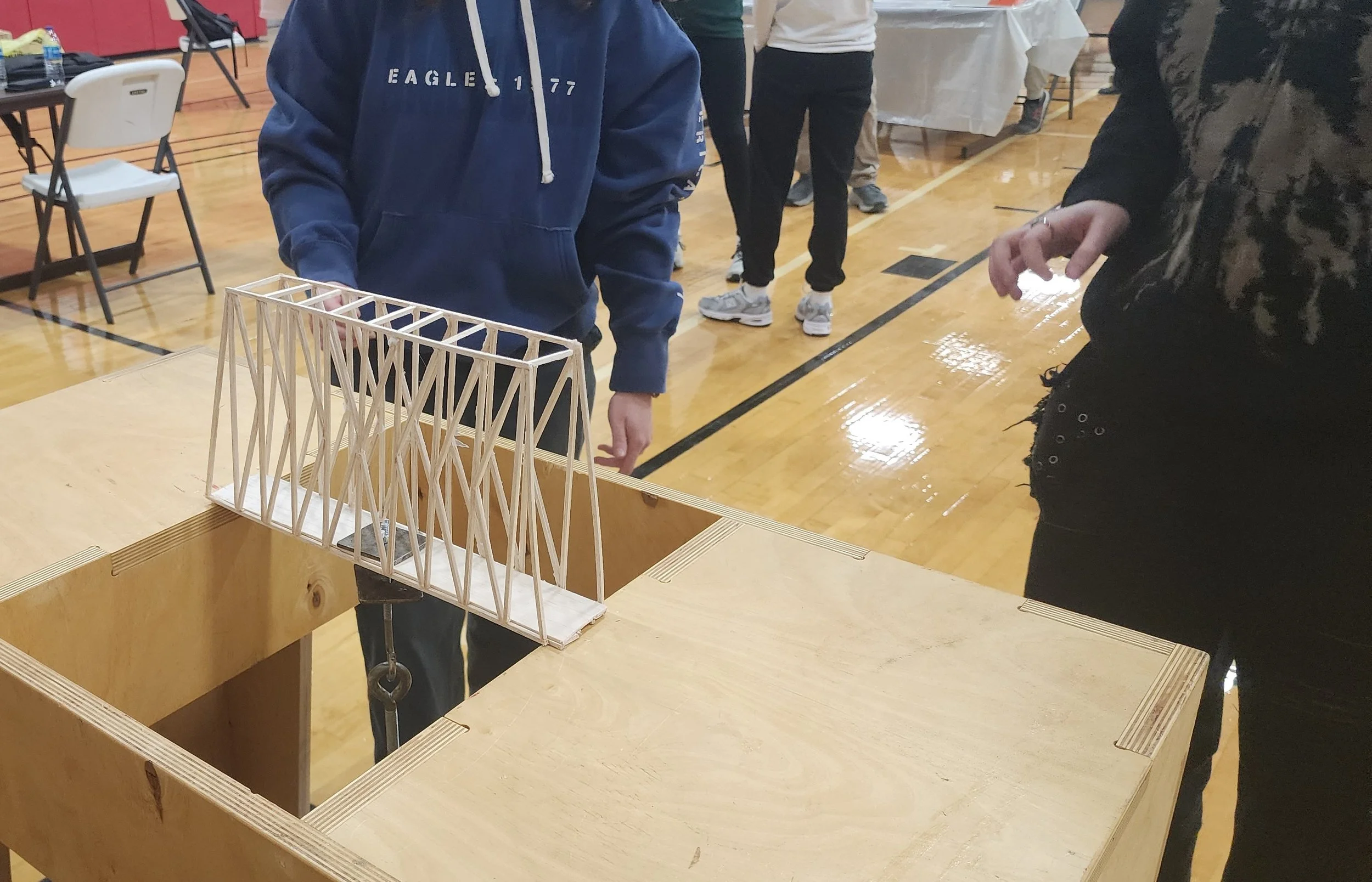 People are gathered around a wooden table in a gymnasium, observing a small wooden bridge model.