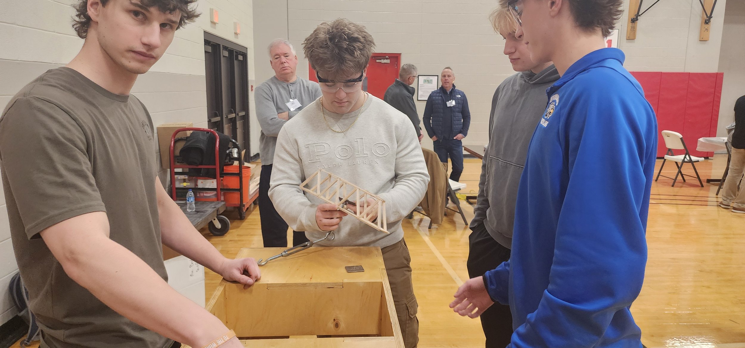 A group of young men examining a wooden bridge model at an indoor event, with two men in the background observing.