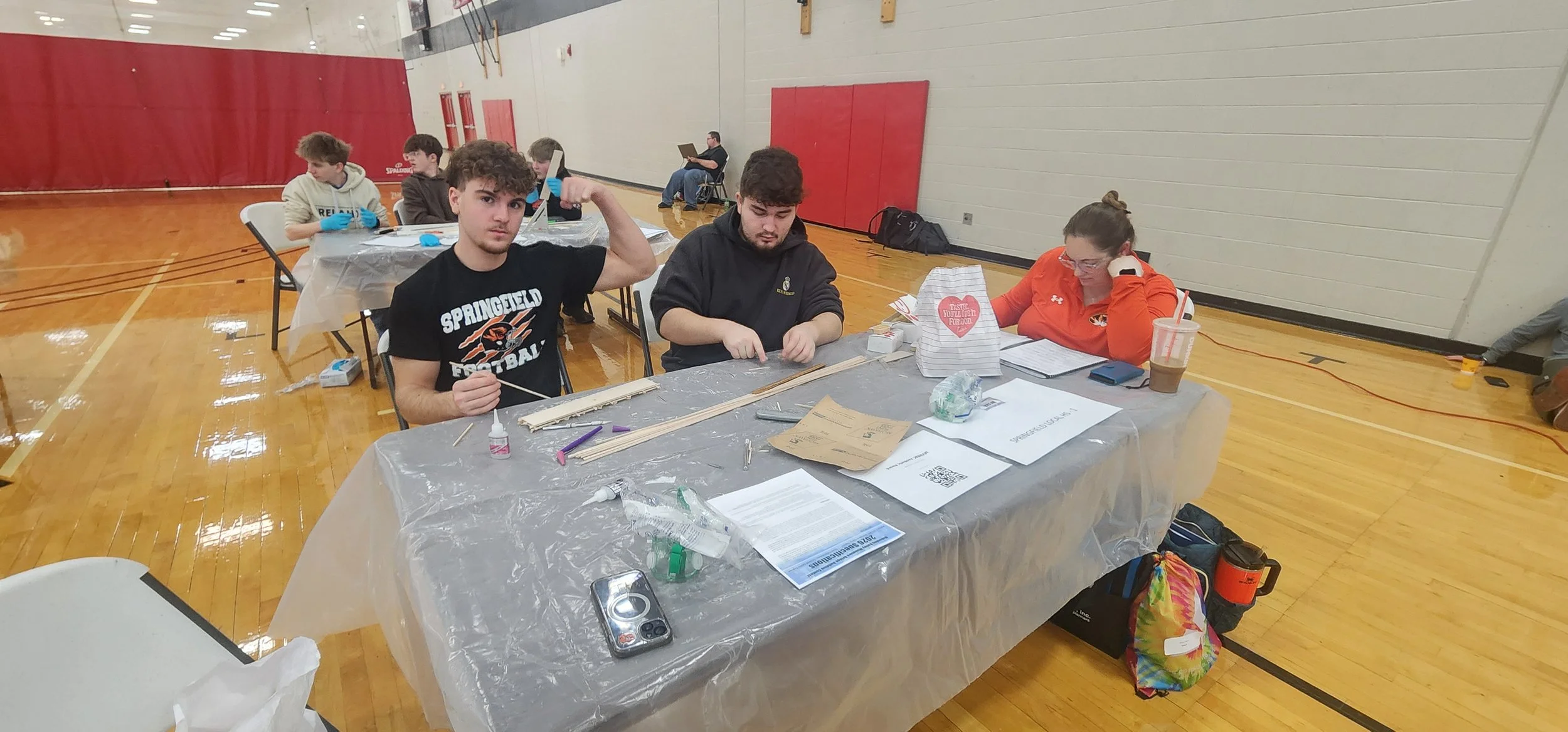 A group of young people sitting at a table in a gymnasium, working on a project involving wooden sticks, glue, and papers. One person is flexing their arm, others are reading or working. There are supplies like water bottles, papers, and a gift bag o