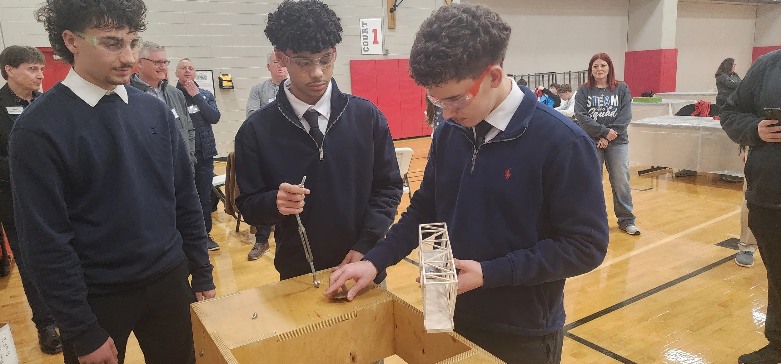 Three young men in safety glasses inspecting a small structural model on a wooden table at a school or community event in a gymnasium.