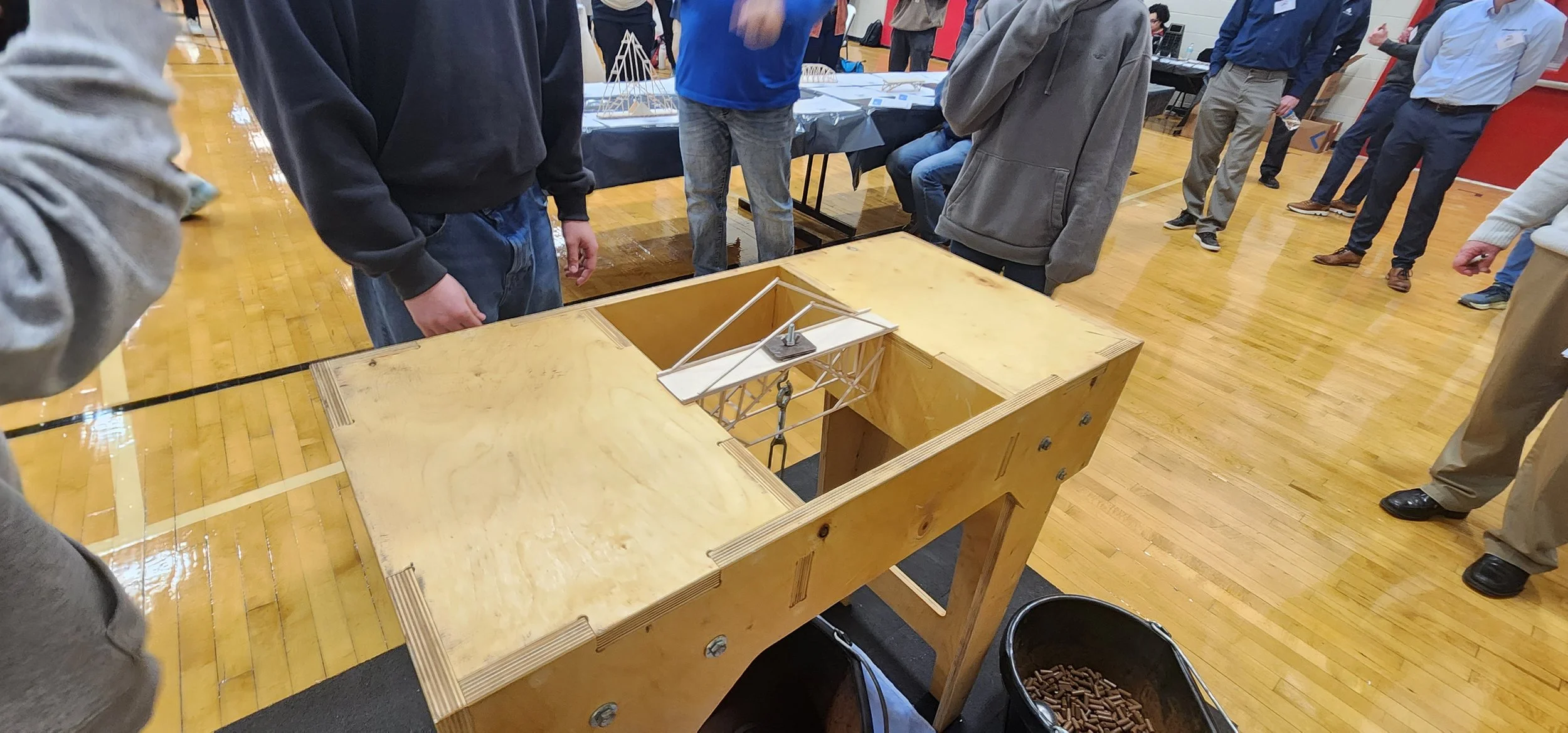 People gathered around a wooden table in a gymnasium, observing a small wooden and metal mechanical model or device on the table.