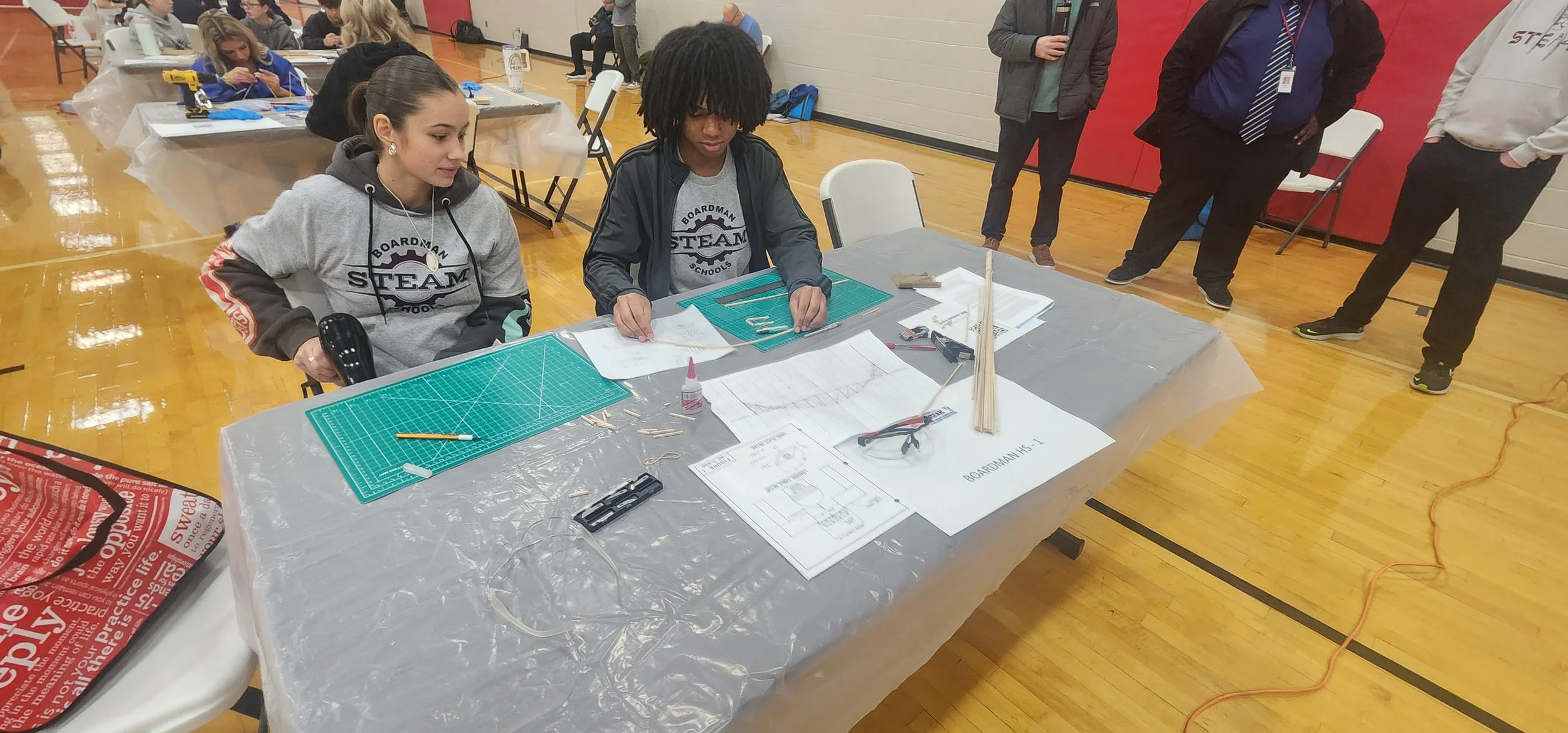 Two students working on robotics projects at a table with tools, plans, and materials during a school event in a gymnasium.