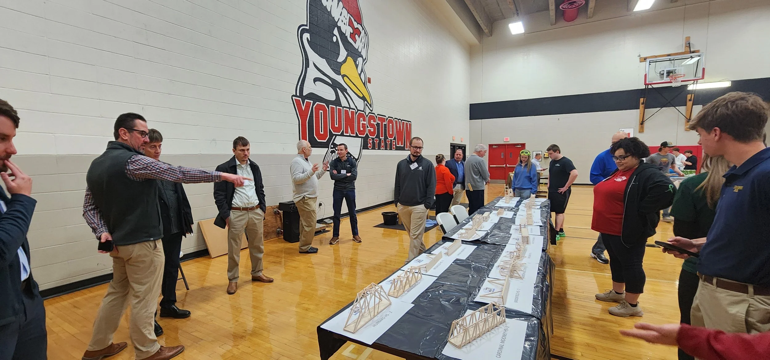 Group of people gathered in a gymnasium at an event, looking at a display of miniature bridge models on tables. Large 'YOUNGSTOWN STATE' sign with a bird logo on the wall in the background.