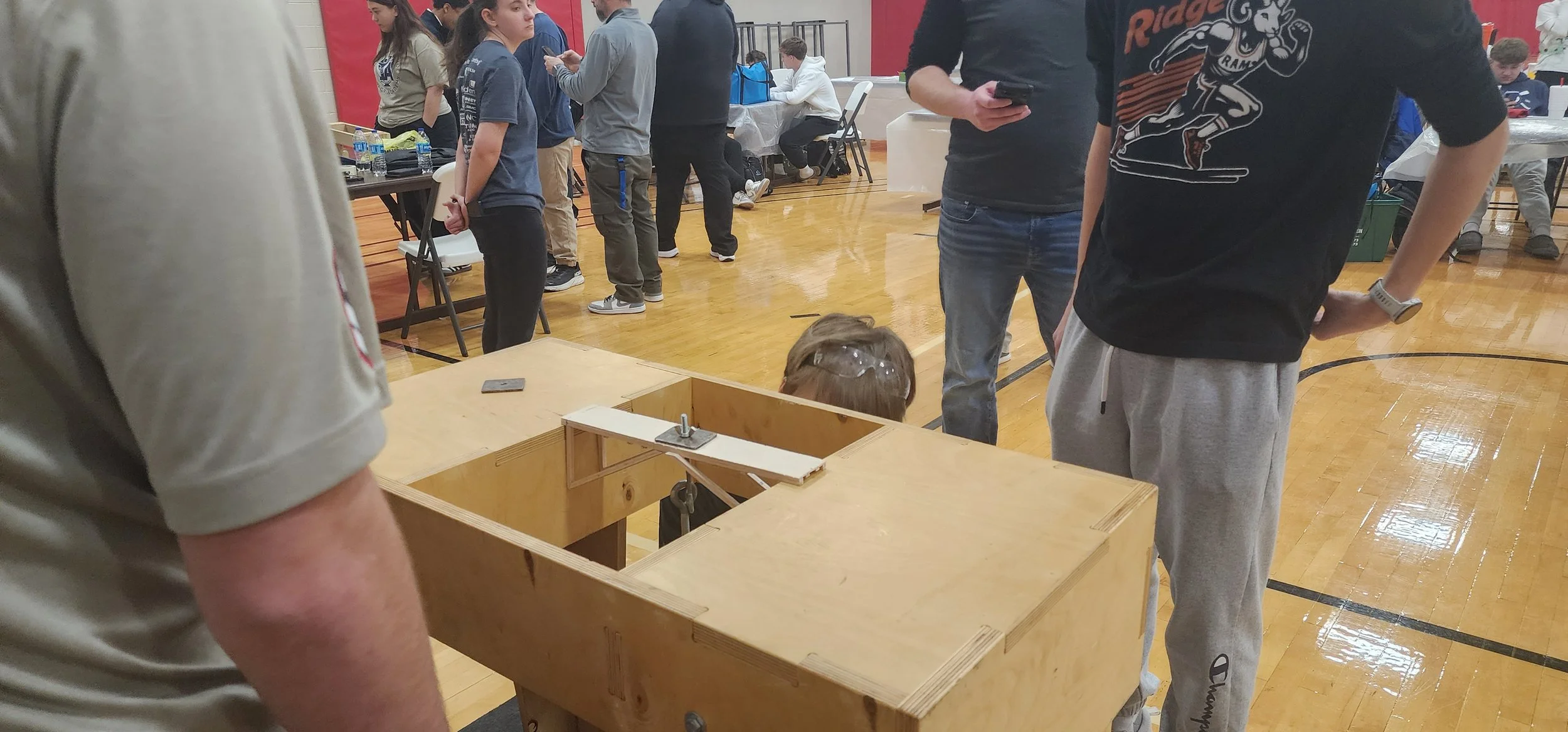 A person is working on a woodworking project in a gymnasium, surrounded by people observing. The project appears to be a wooden box with some parts attached, and a child's head is visible inside the box, with safety glasses on.