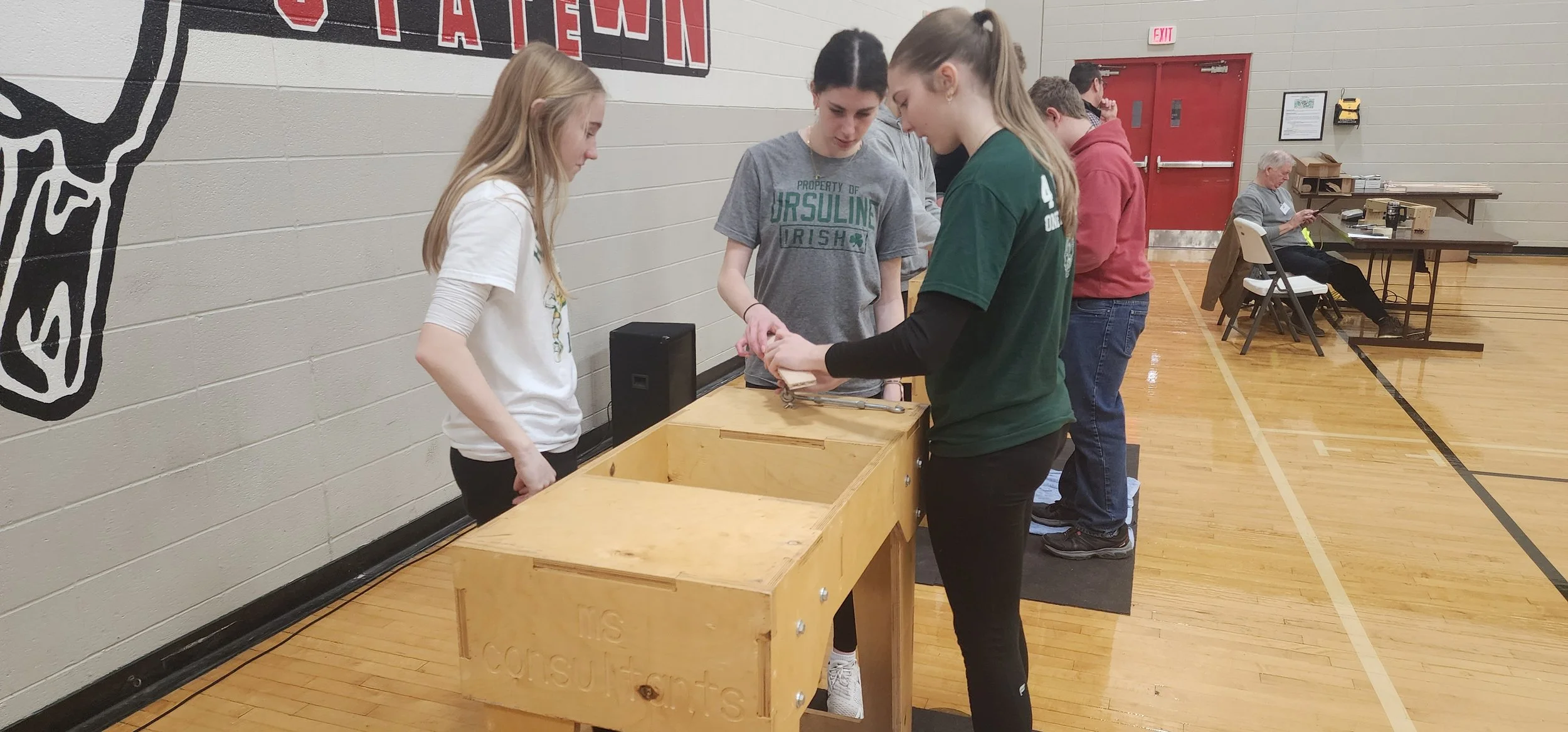 Young people in a gymnasium voting or participating in a poll, with a woman sitting at a table in the background.