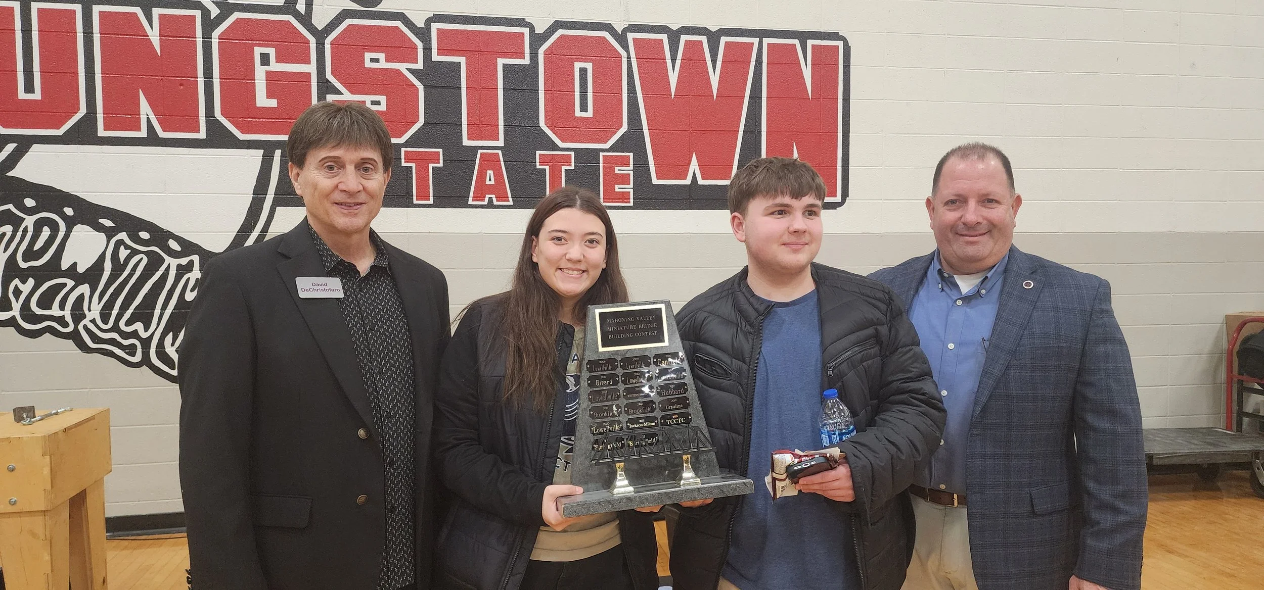 Group of four people standing together indoors with a mural on the wall behind them that says 'Ingh Town State'. The woman in the center is holding a trophy for the Mahoning Valley Miniature Bridge Building Contest. The woman on the left and the man 