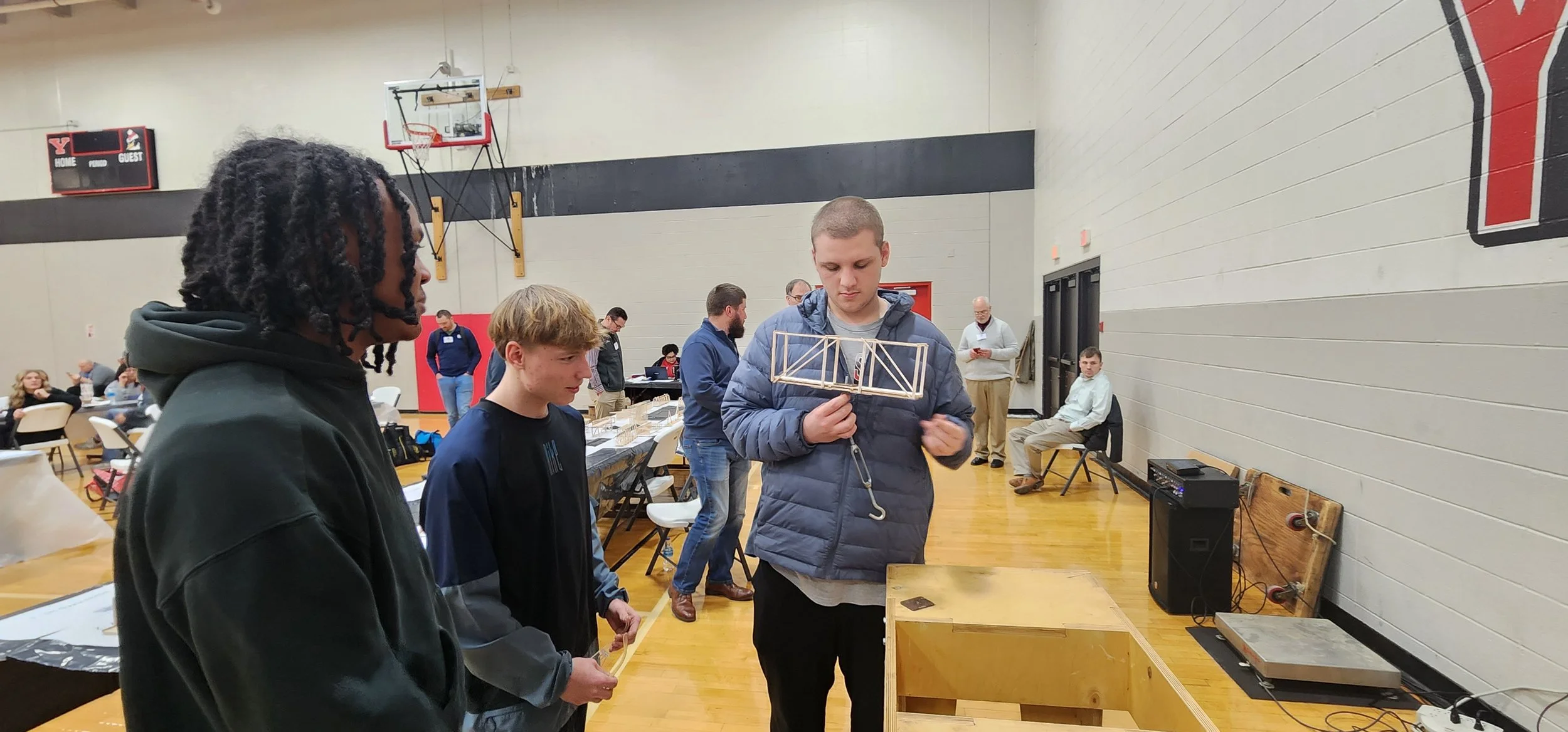 A group of young men in a gymnasium observing a model bridge made of wooden sticks held by one of the men. There are tables, chairs, and other people in the background, some standing and some sitting.