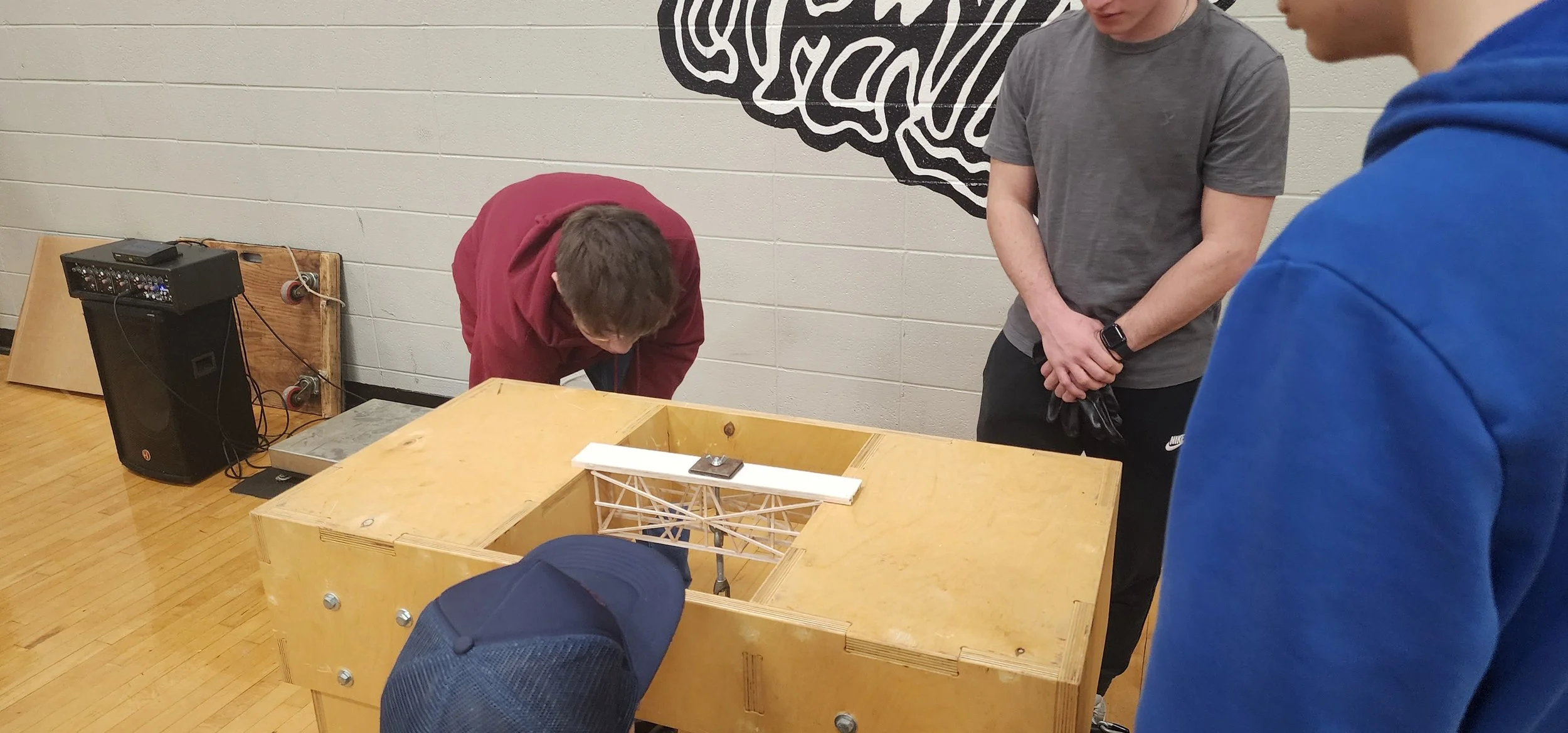 Group of people gathered around a wooden table with a structural model and a small bridge, indoors with a large black and white mural on the wall.