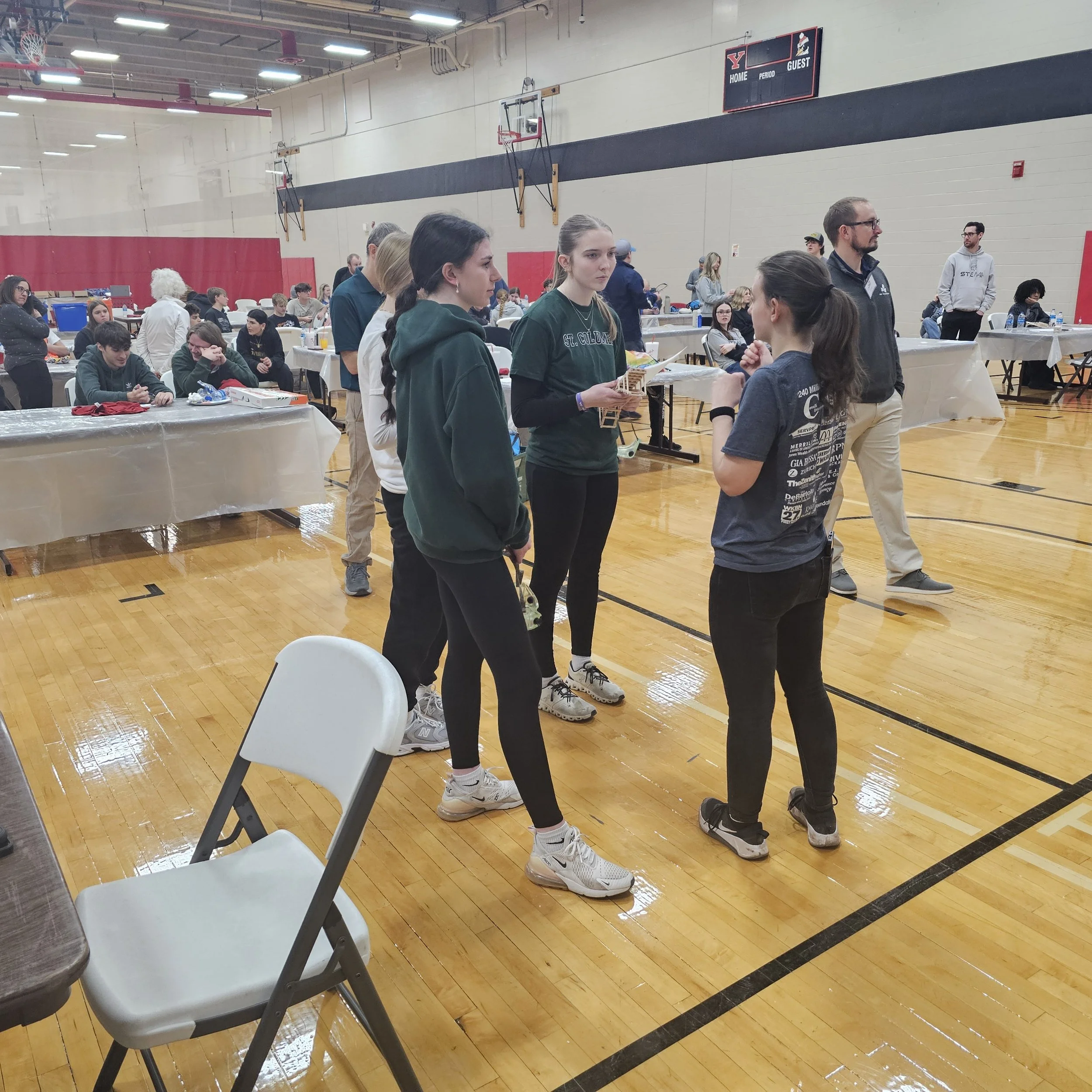 A group of young people gathered in a gymnasium, standing and talking near tables covered with tablecloths. Some are seated while others are standing, with water bottles and various items on the tables. The gym has a basketball hoop, a scoreboard, an