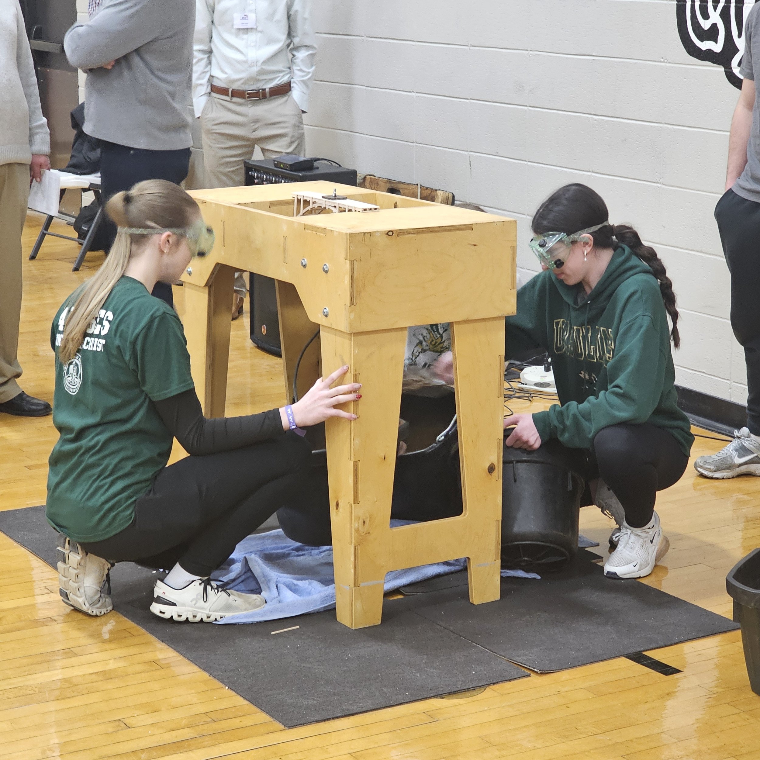 People sitting on the floor and working on a wooden school project, with some standing around, in a gymnasium with a white wall and wooden floor.