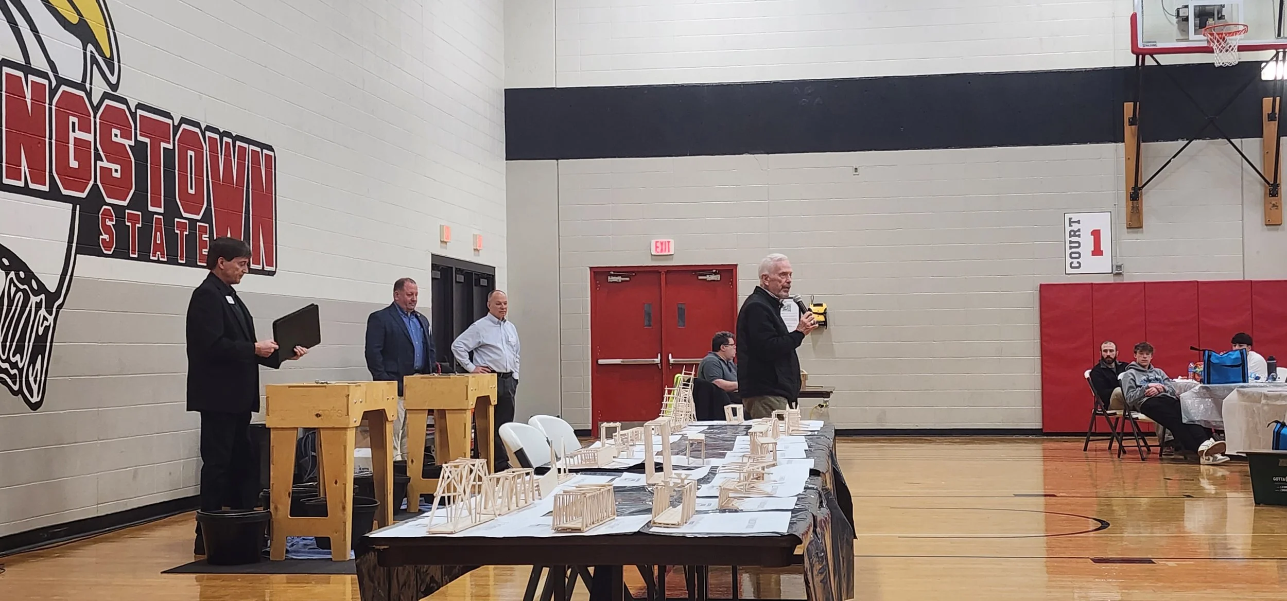 A man speaking into a microphone at an indoor event in a gymnasium, with a table displaying wooden architectural models and several seated and standing people.