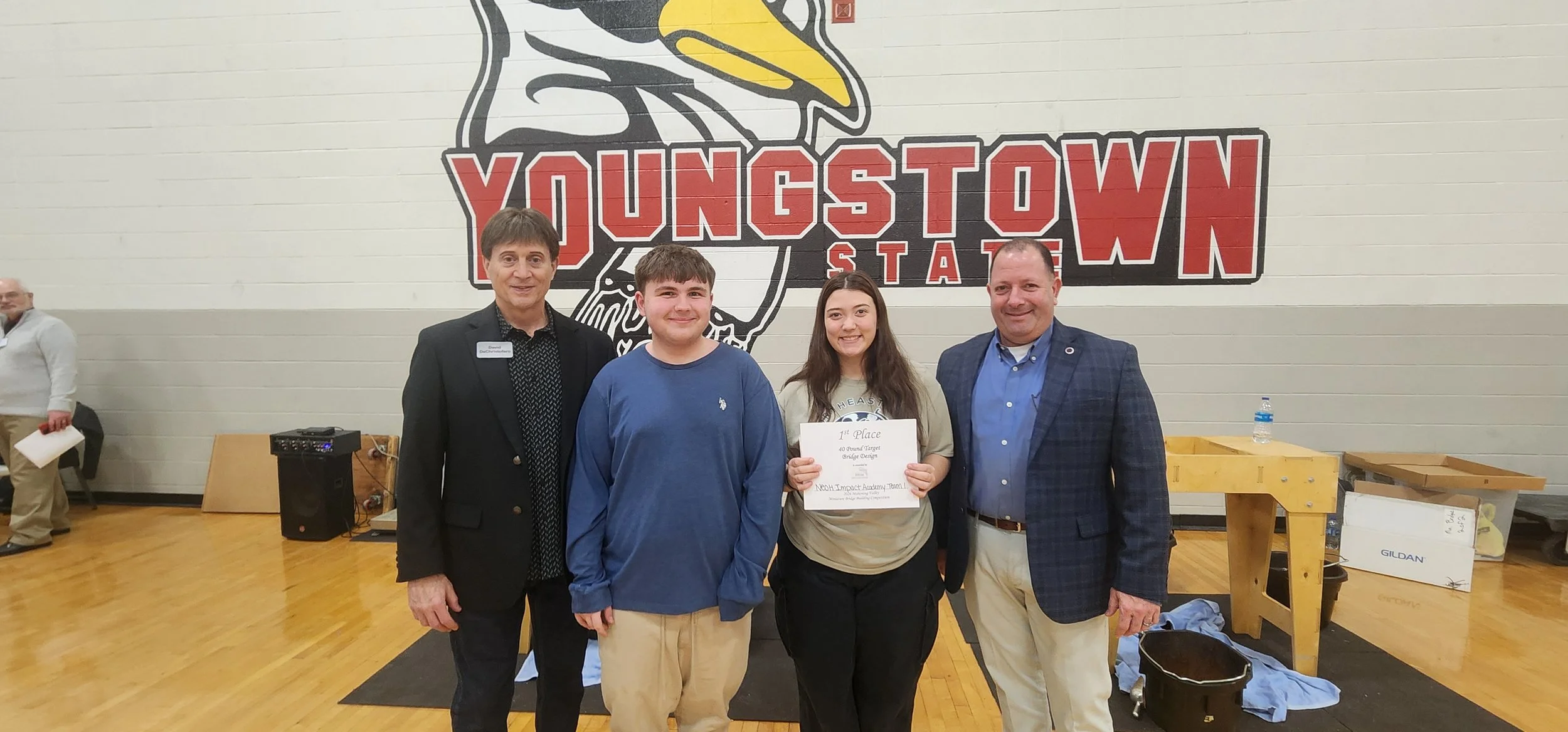 Group of four people standing in front of a large 'YOUNGSTOWN' and a bird graphic on a gymnasium wall, with a girl holding a certificate that says '1st Place,' at an indoor awards event.