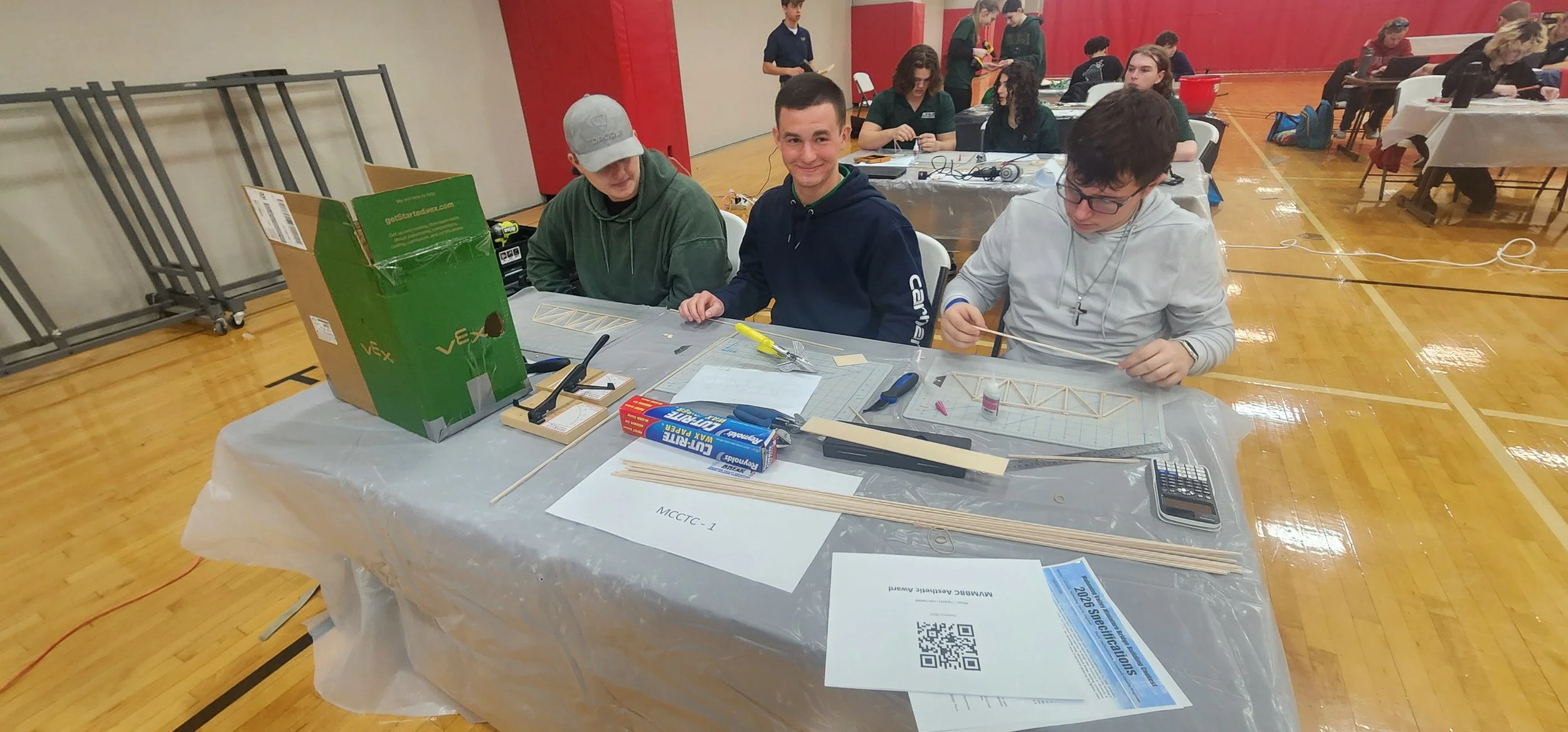 Four young men sit at a table working on a model aircraft or bridge, surrounded by tools, materials, and instructions, with other groups in the background working on projects in a gymnasium.