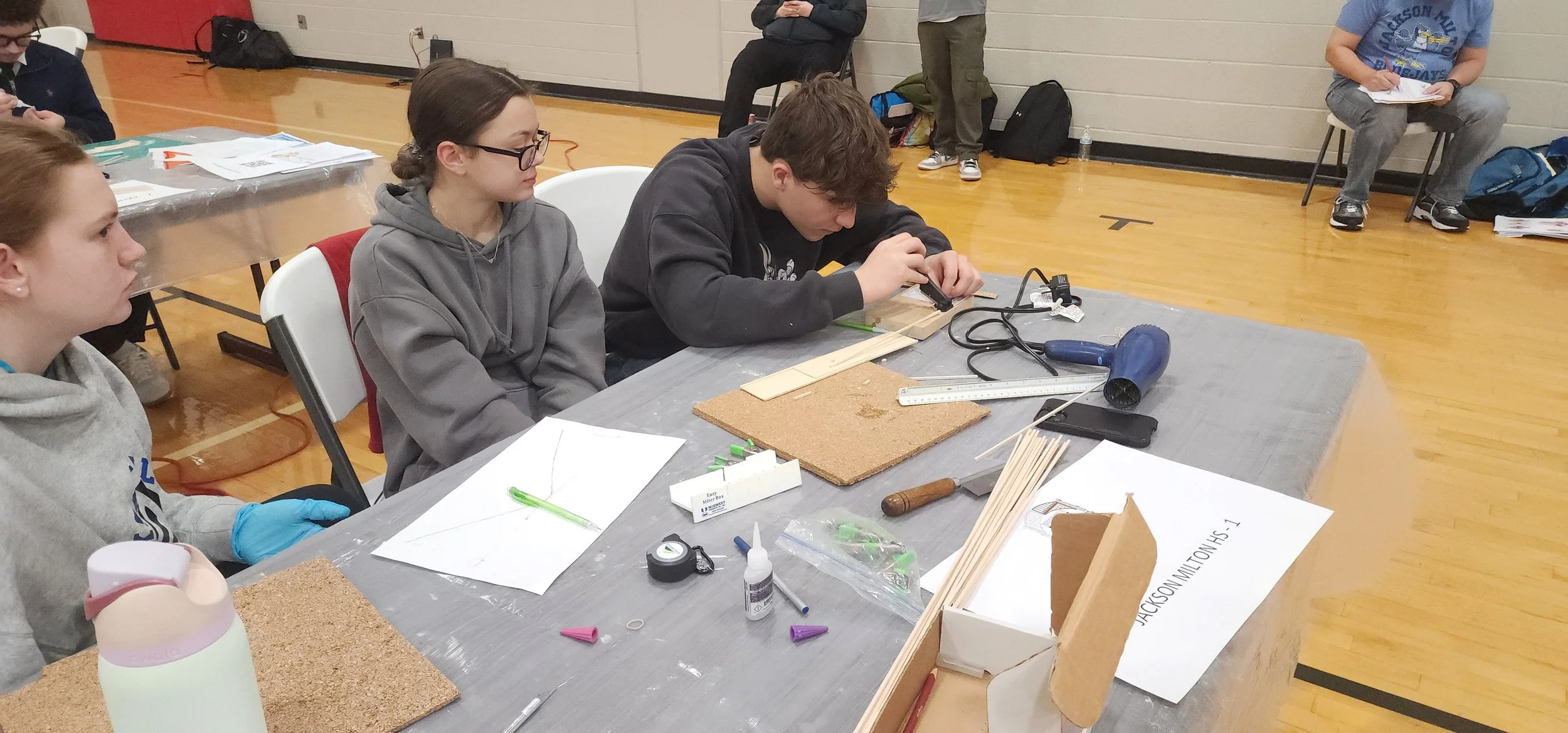 Students working on a science project at a table with various materials such as cork, sticks, tools, and papers in a school gymnasium.
