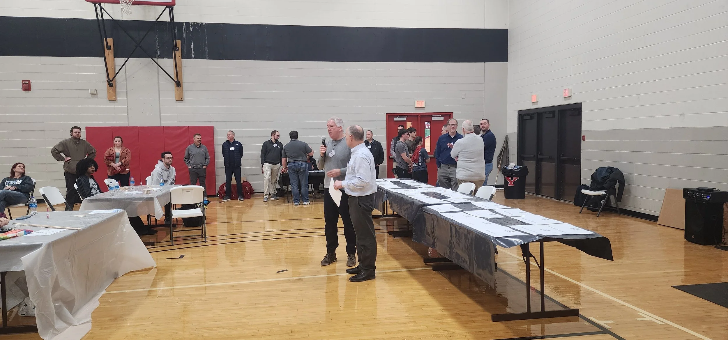 People gathered in a gymnasium for an event, with some seated at tables and others standing and talking, including a man speaking into a microphone.