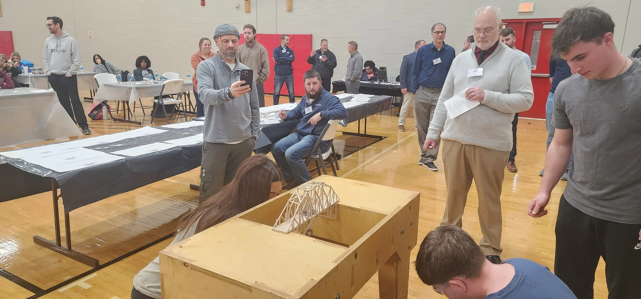 People gathered in a gymnasium observing a wooden model bridge on a table.