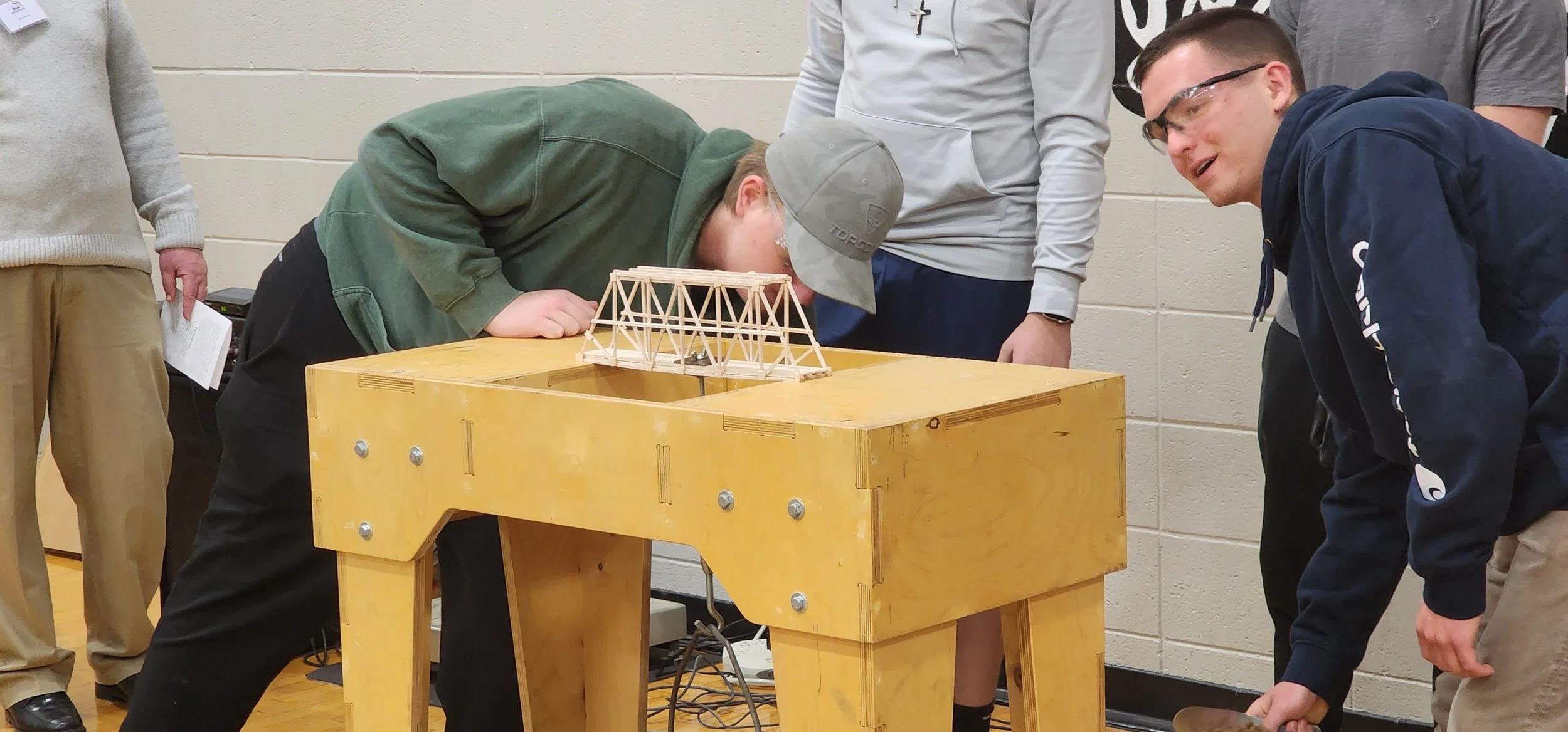 Group of young people gathered around a wooden table with a small wooden bridge model, one boy leaning over and looking closely, others observing and smiling.