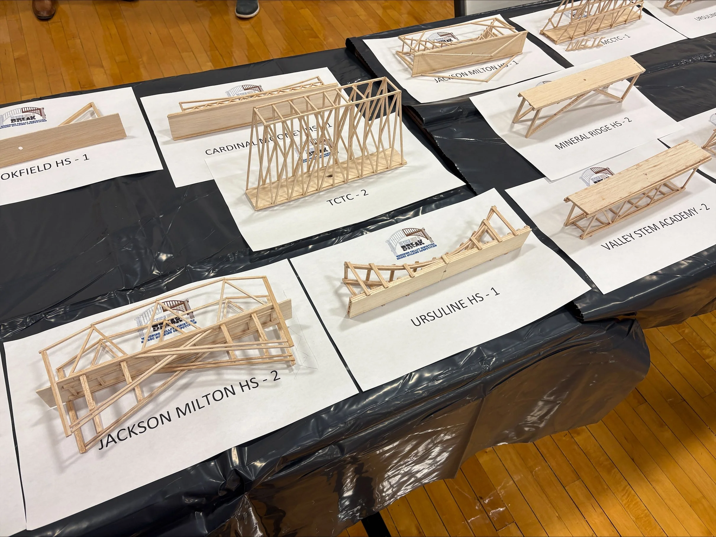 Multiple miniature wooden architectural models displayed on a table, each labeled with names such as Jackson Milton HS, Cardinham Elementary, Mineral Ridge HS, Ursuline HS, and Valley STEM Academy, on white paper sheets on a black plastic-covered tab