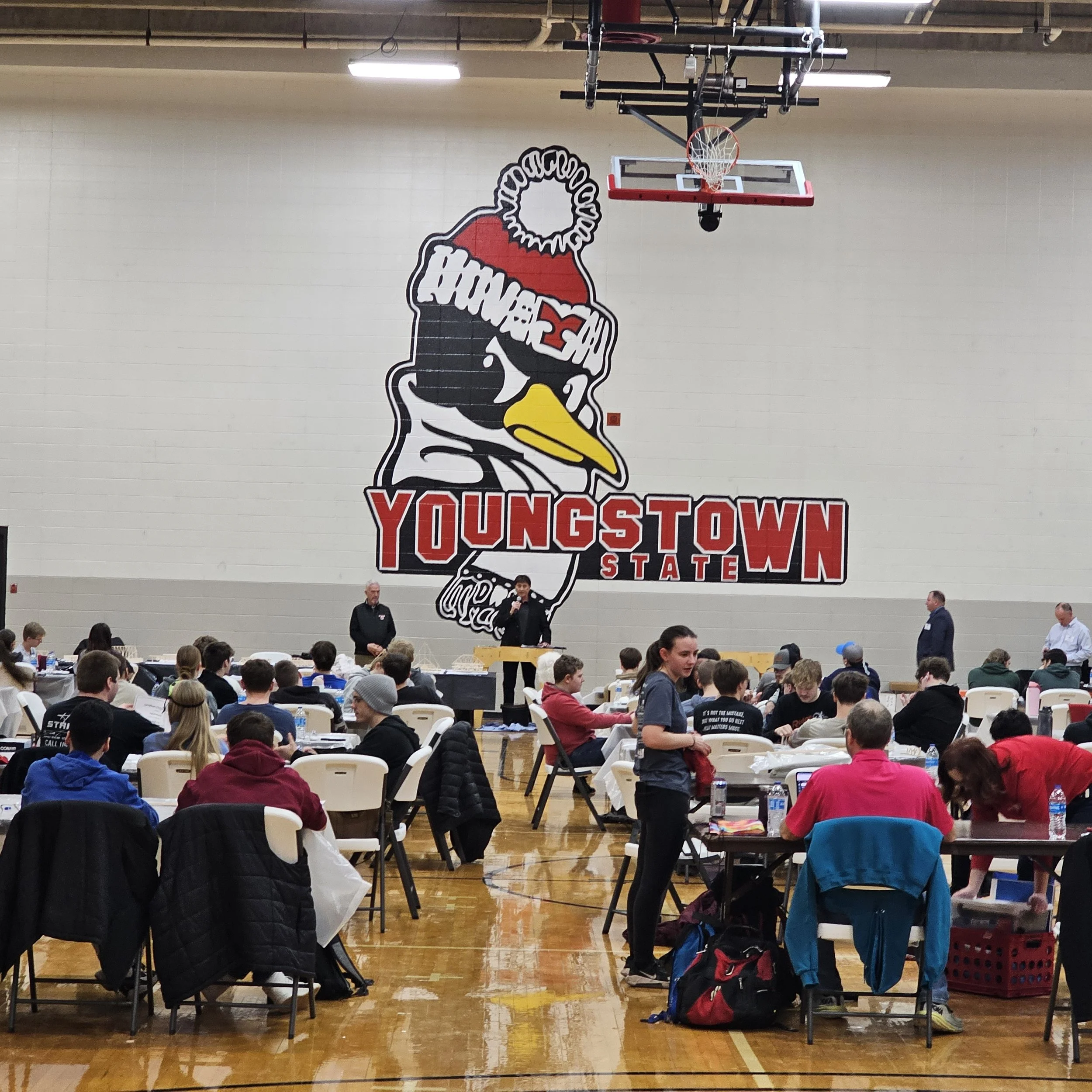 A gymnasium filled with people attending an event, with a large mural of a cartoon duck wearing a hoodie and cap on the wall. The basketball hoop is overhead, and people are seated at tables in front.
