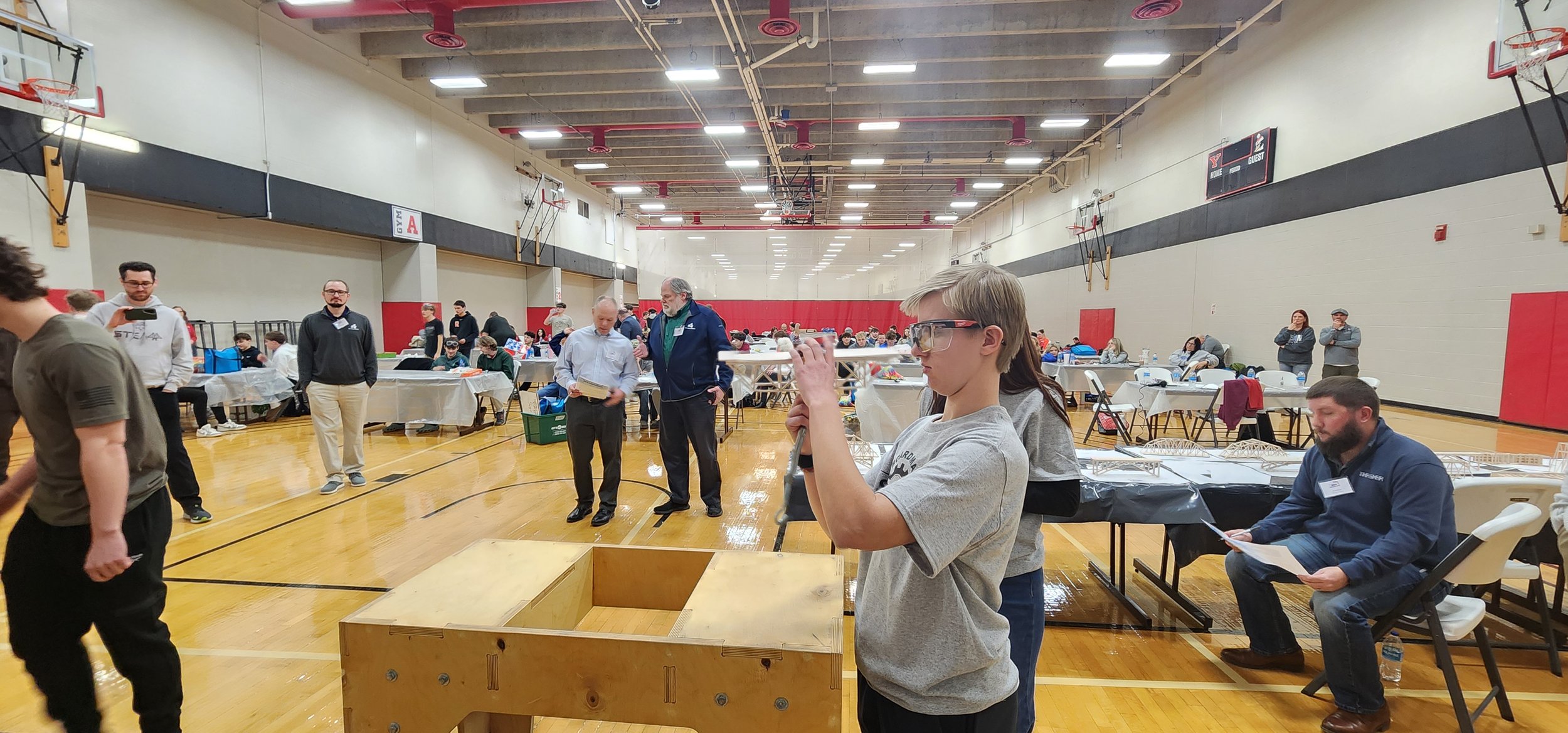 People gathered in a gymnasium for a robotics event, with tables displaying models and attendees engaging in conversation and activities.
