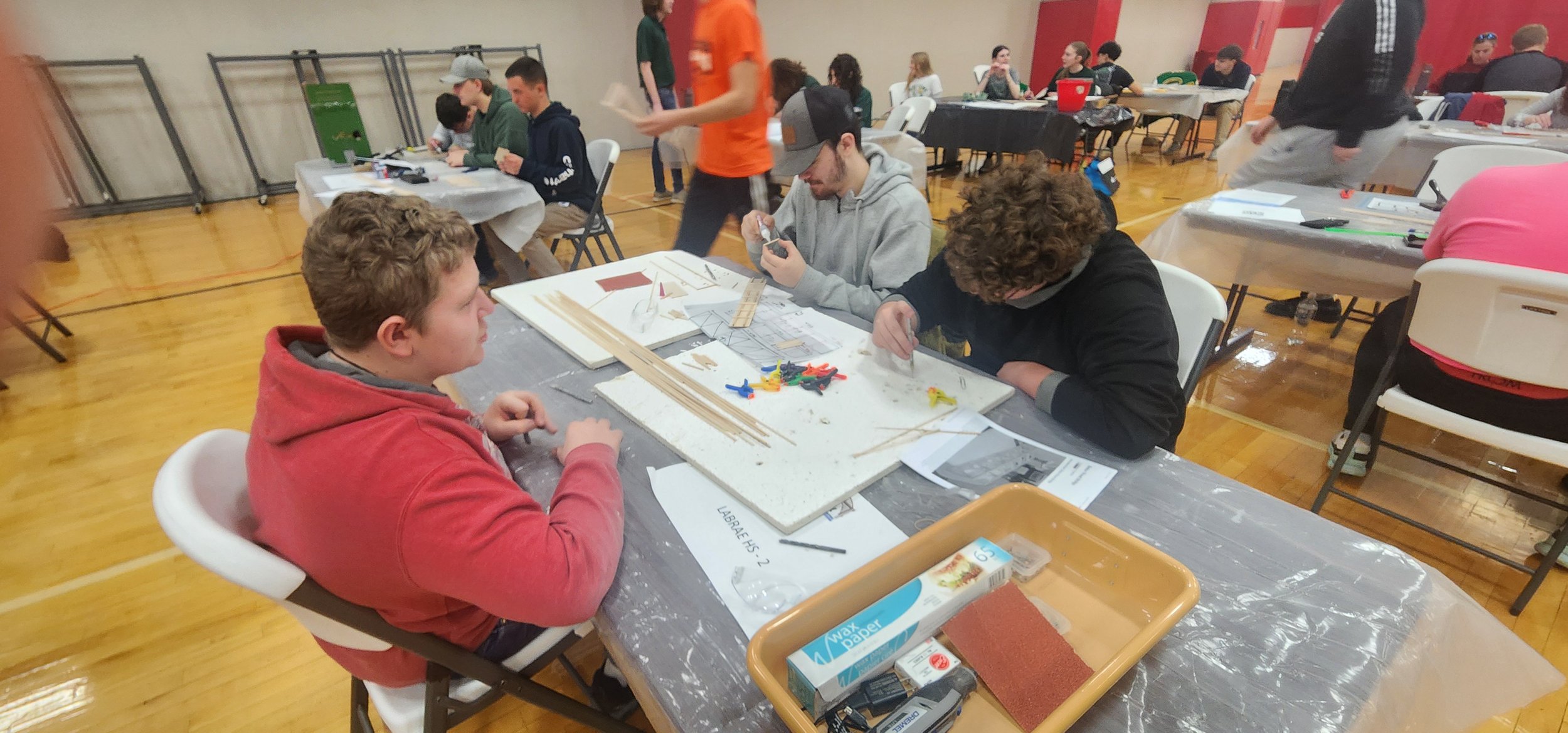 Young people working on a model or project at a table with various tools and materials in a large indoor space.