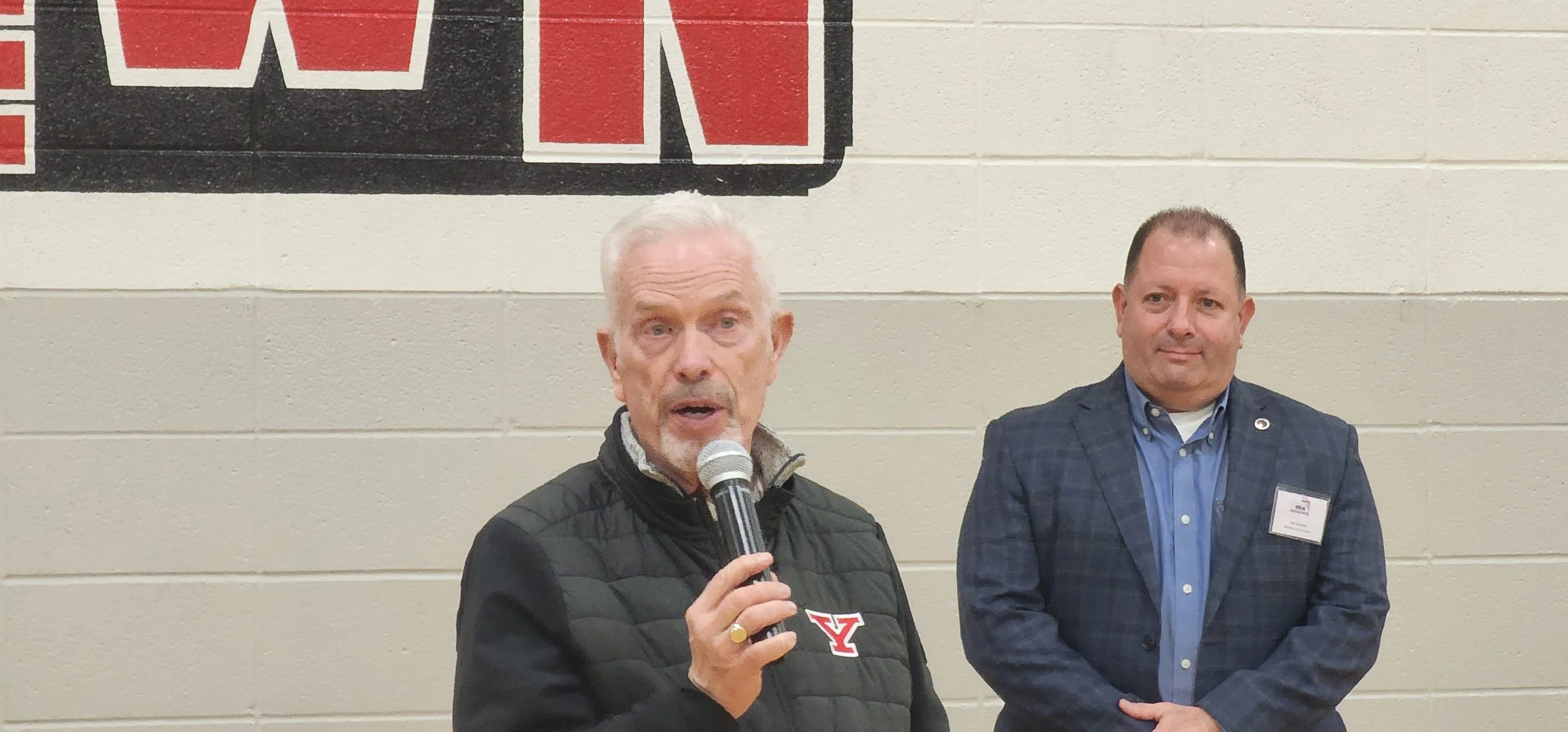 An elderly man with white hair speaking into a microphone, wearing a black jacket with a Yale logo, standing next to a man in a blue checkered blazer with a name tag, both in front of a beige brick wall with a partially visible banner.
