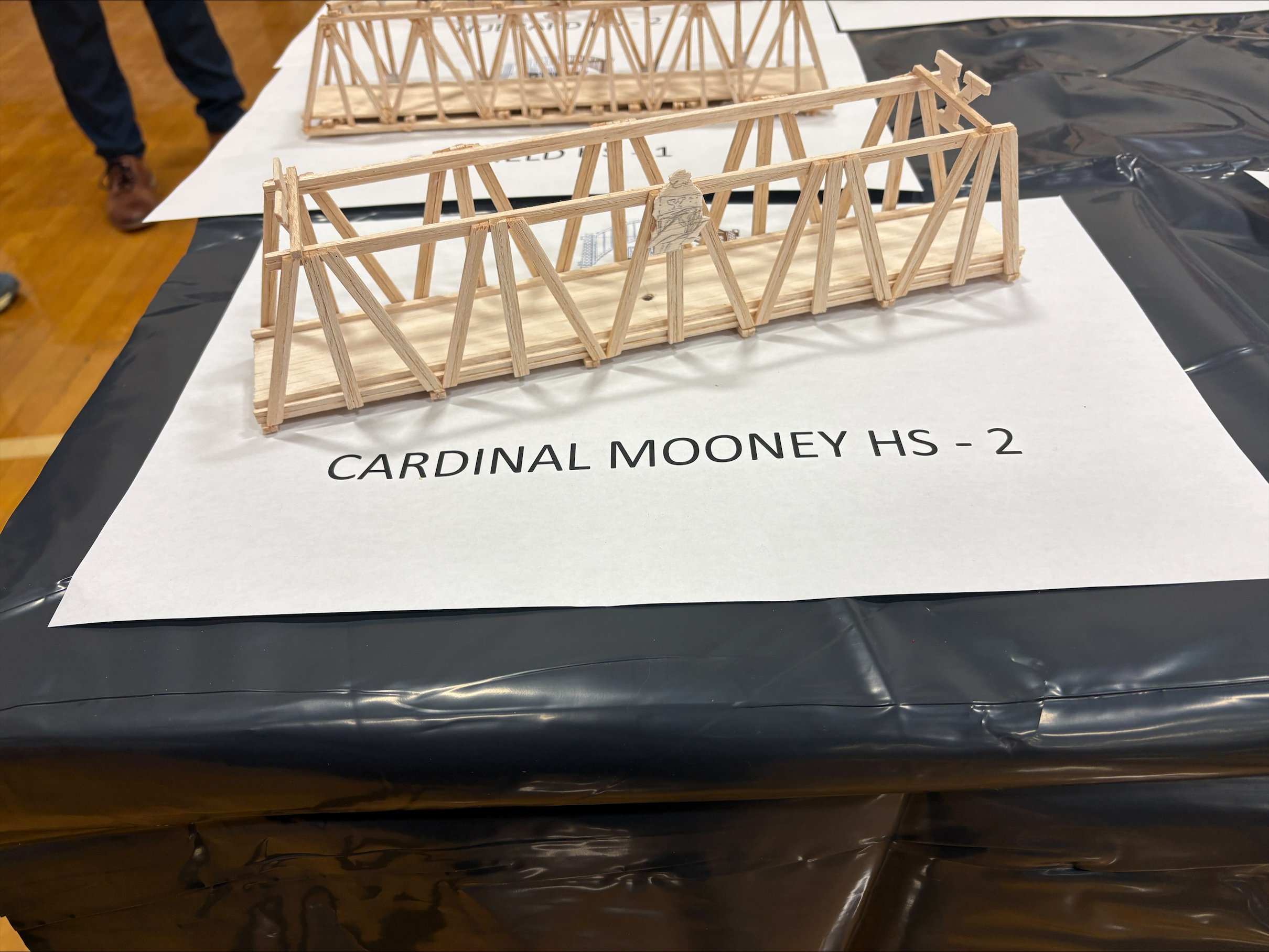 Two detailed model bridges made of wooden sticks are displayed on a table with a white surface, labeled "CARDINAL MOONEY HS - 2". The background includes a person wearing dark pants and brown shoes, standing on a wooden floor.