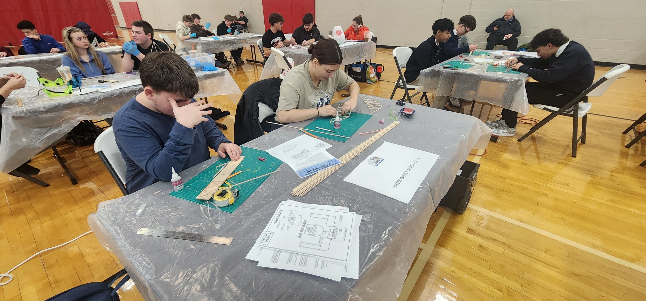 Group of students working on engineering projects in a gymnasium classroom, with various tools and sketches on the table.