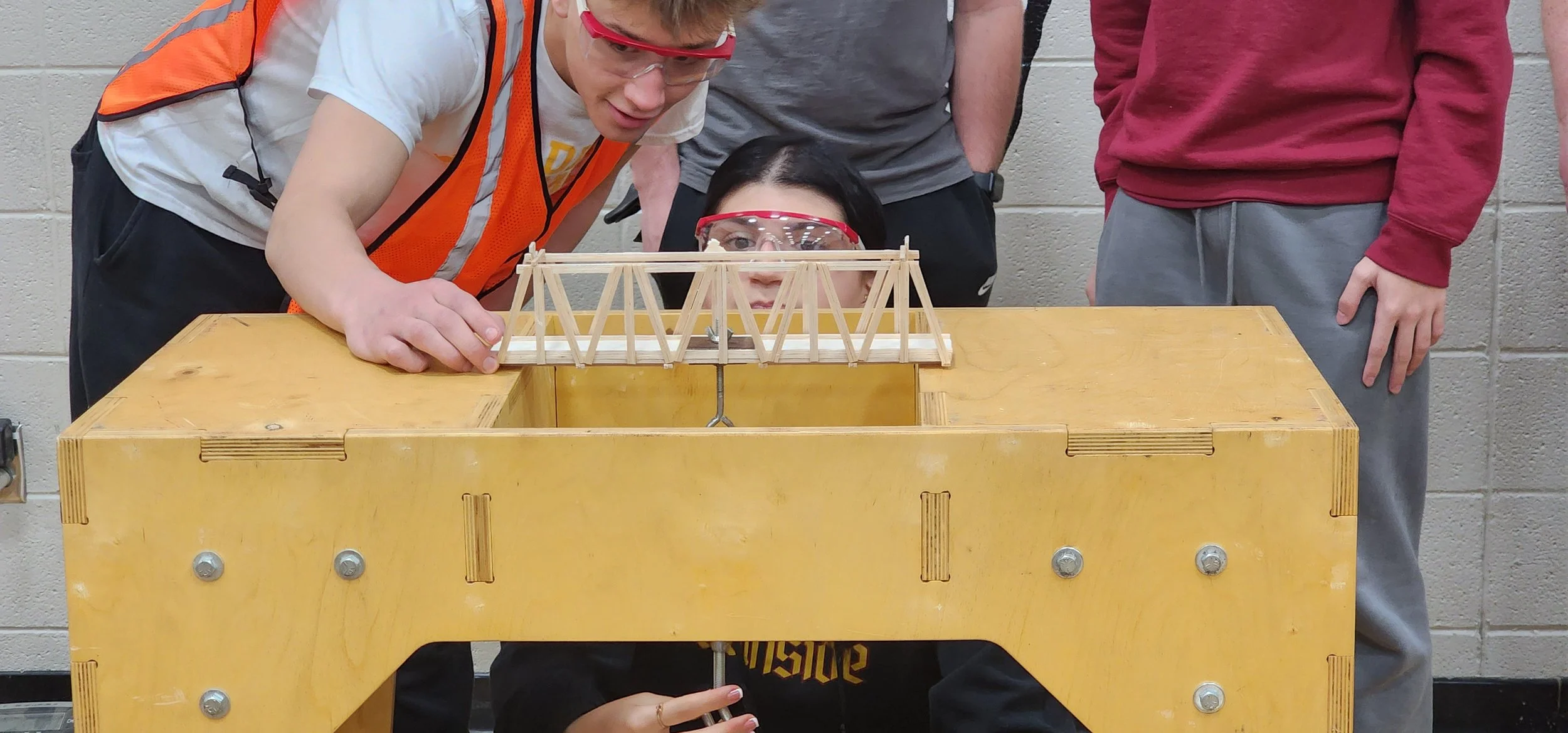 Group of students testing a small bridge model made of sticks in a classroom lab.