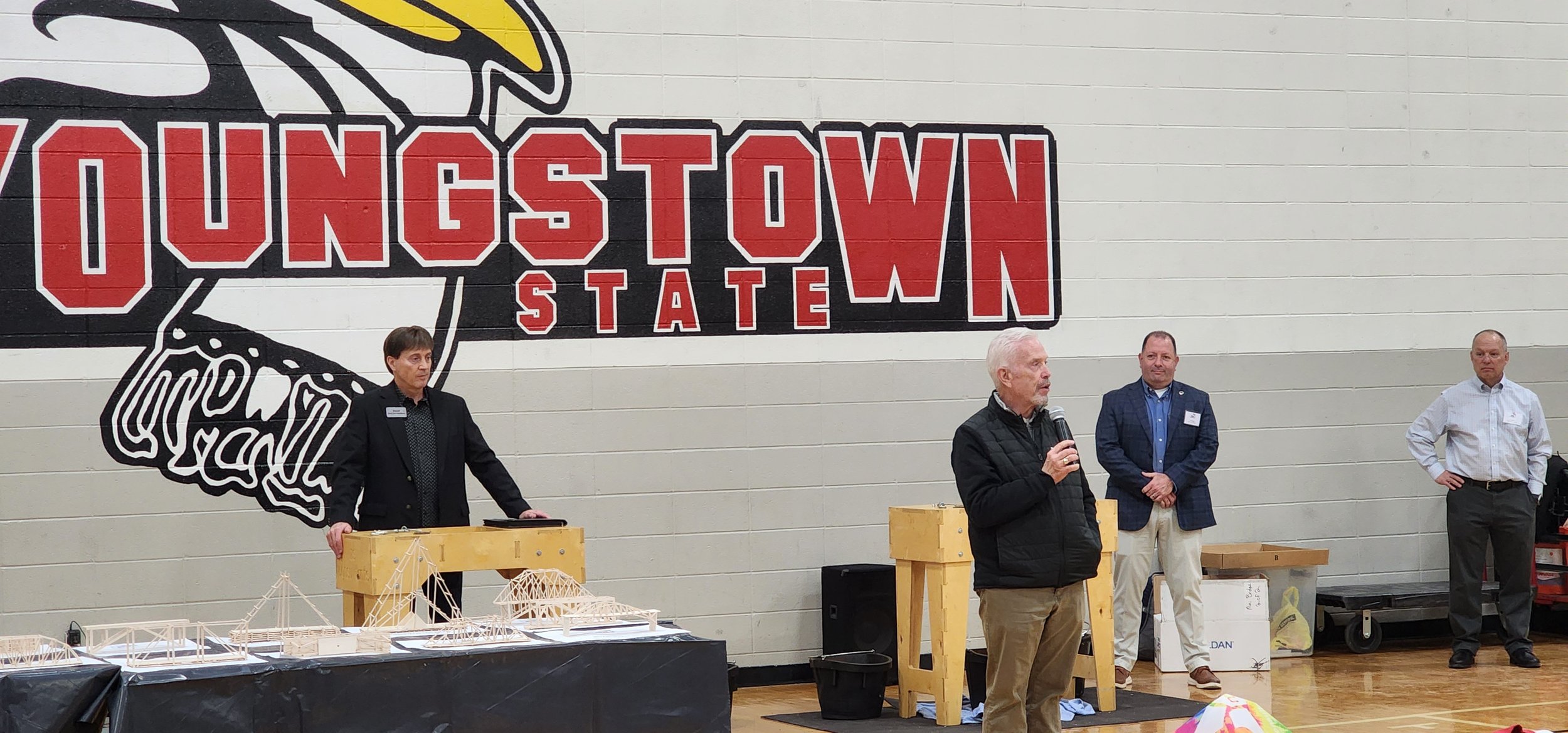 A group of four men standing in front of a large wall with a mural that reads 'LIONESTOWN' and a tiger illustration. One man is speaking into a microphone while others listen. There are tables with models and materials in front of them.