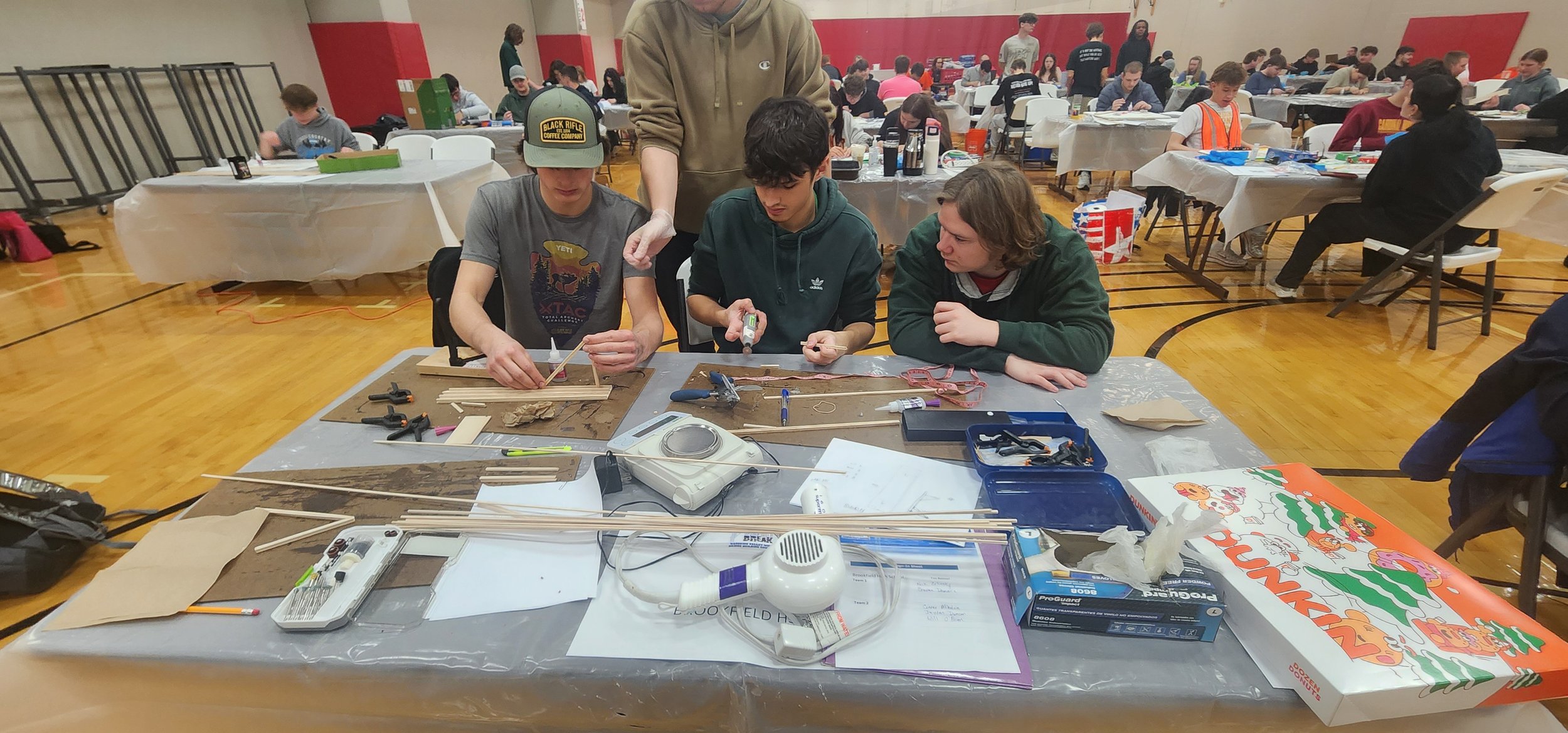 Three young men working on a woodworking project at a table covered with tools and materials in a large indoor gymnasium filled with people.
