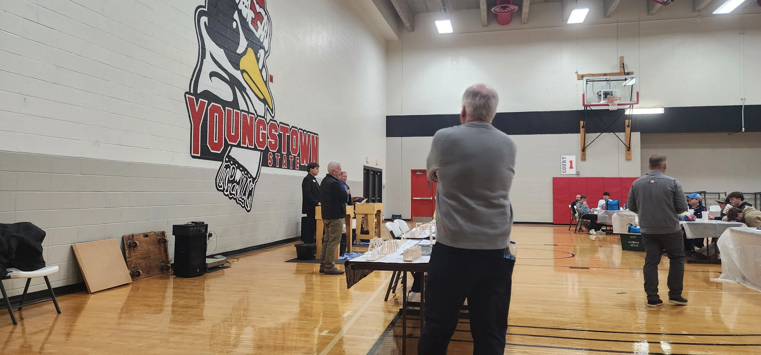 People gathered in a gymnasium for an event, with some standing and some seated at tables. The gym has a large logo on the wall that says "YOUNGSTOWN STATE," featuring a stylized bird mascot. There are tables with items and a speaker area with a podi
