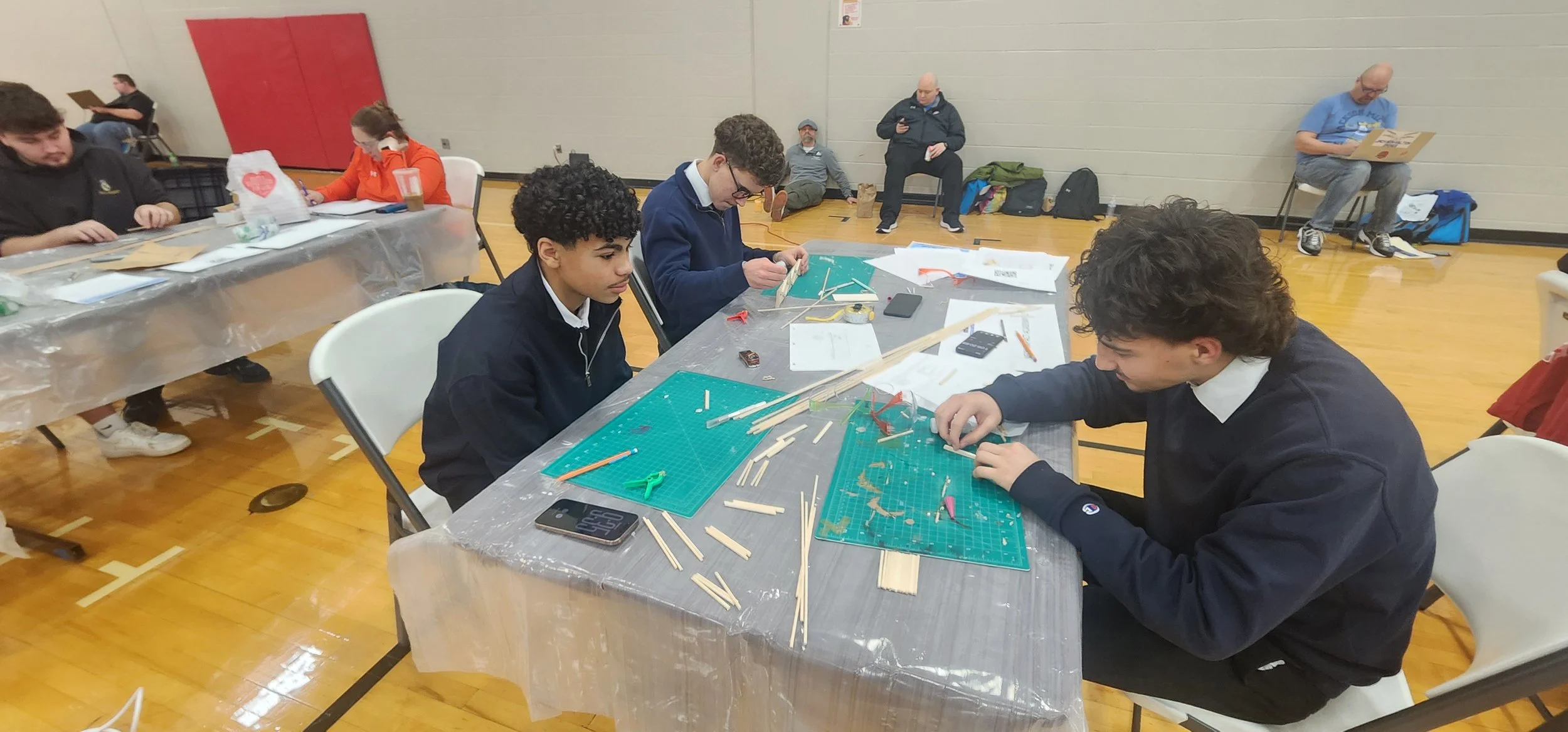 Young students working on a hands-on project with wooden sticks at a table covered with a green cutting mat, tools, and papers, in a gymnasium setting.