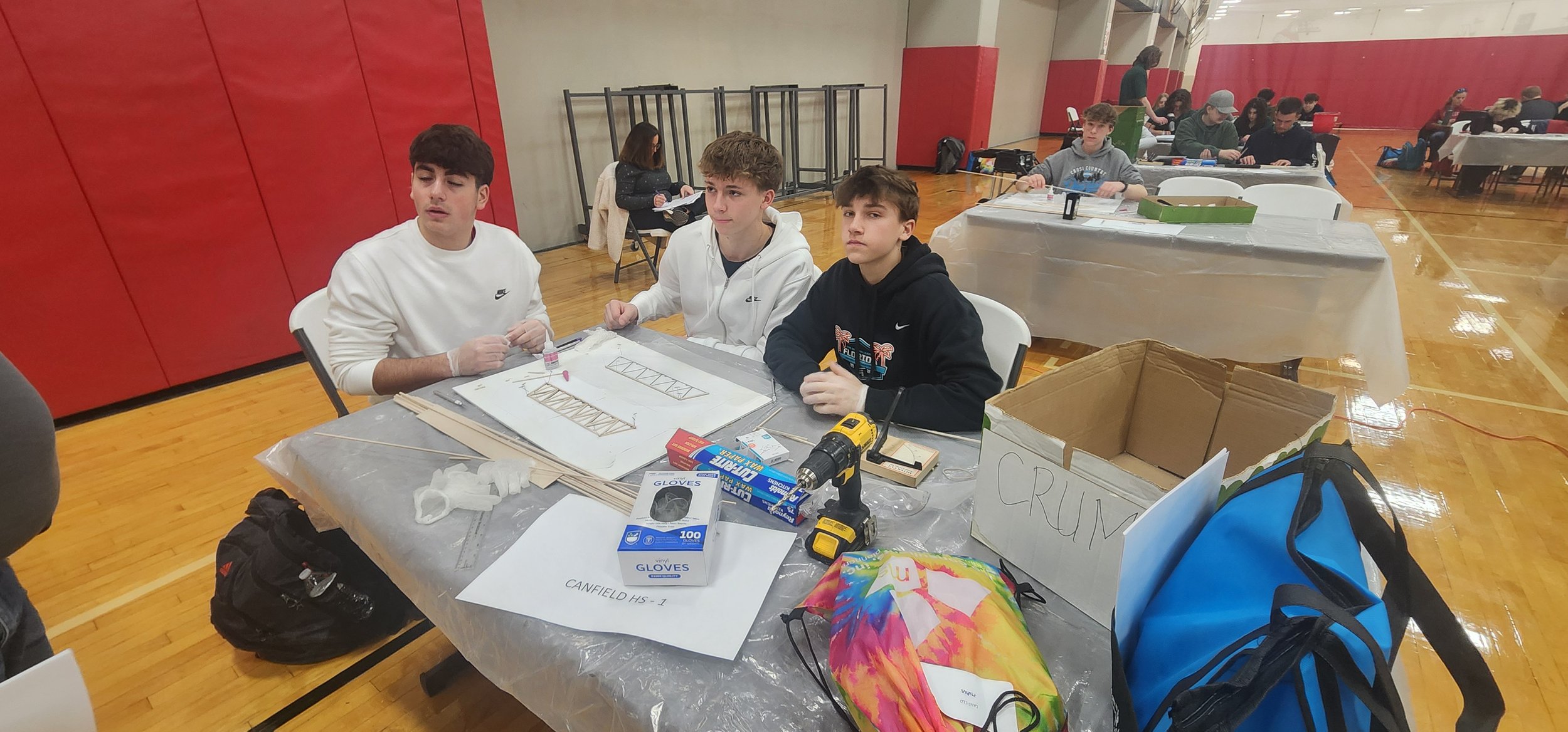 Three boys sitting at a table working on a model construction project in a gymnasium. The table has various tools, materials, and supplies including a drill, gloves, and wooden sticks. Other people are working at tables in the background.