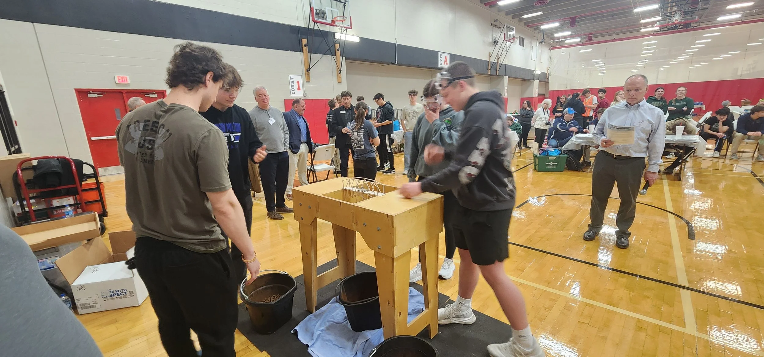 Young people participating in a science or engineering activity in a gymnasium, with others observing in the background.